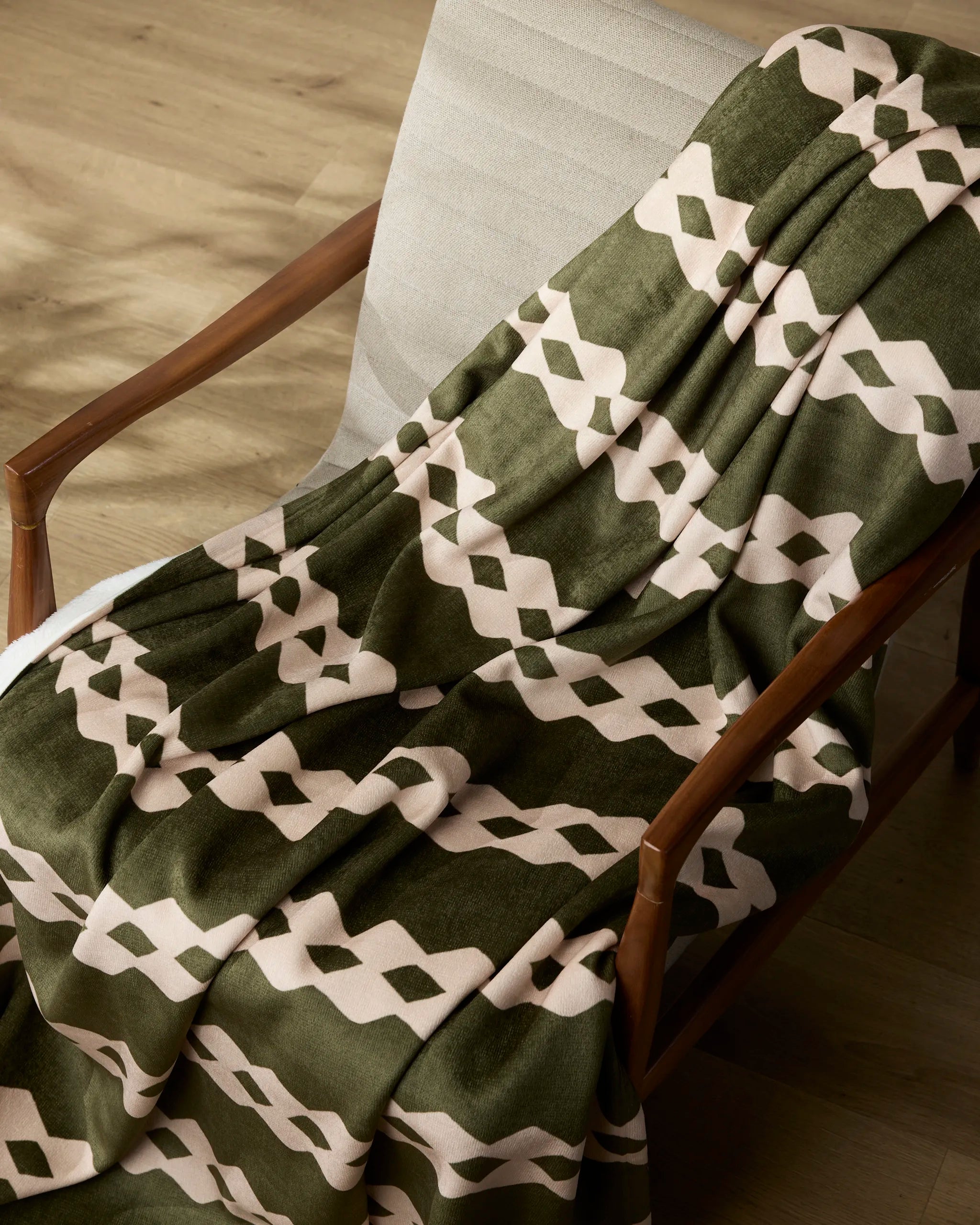 The Geometry Olive Orchard throw blanket, featuring a green and beige geometric pattern, is draped over a light cushioned chair with wooden arms on a wooden floor.