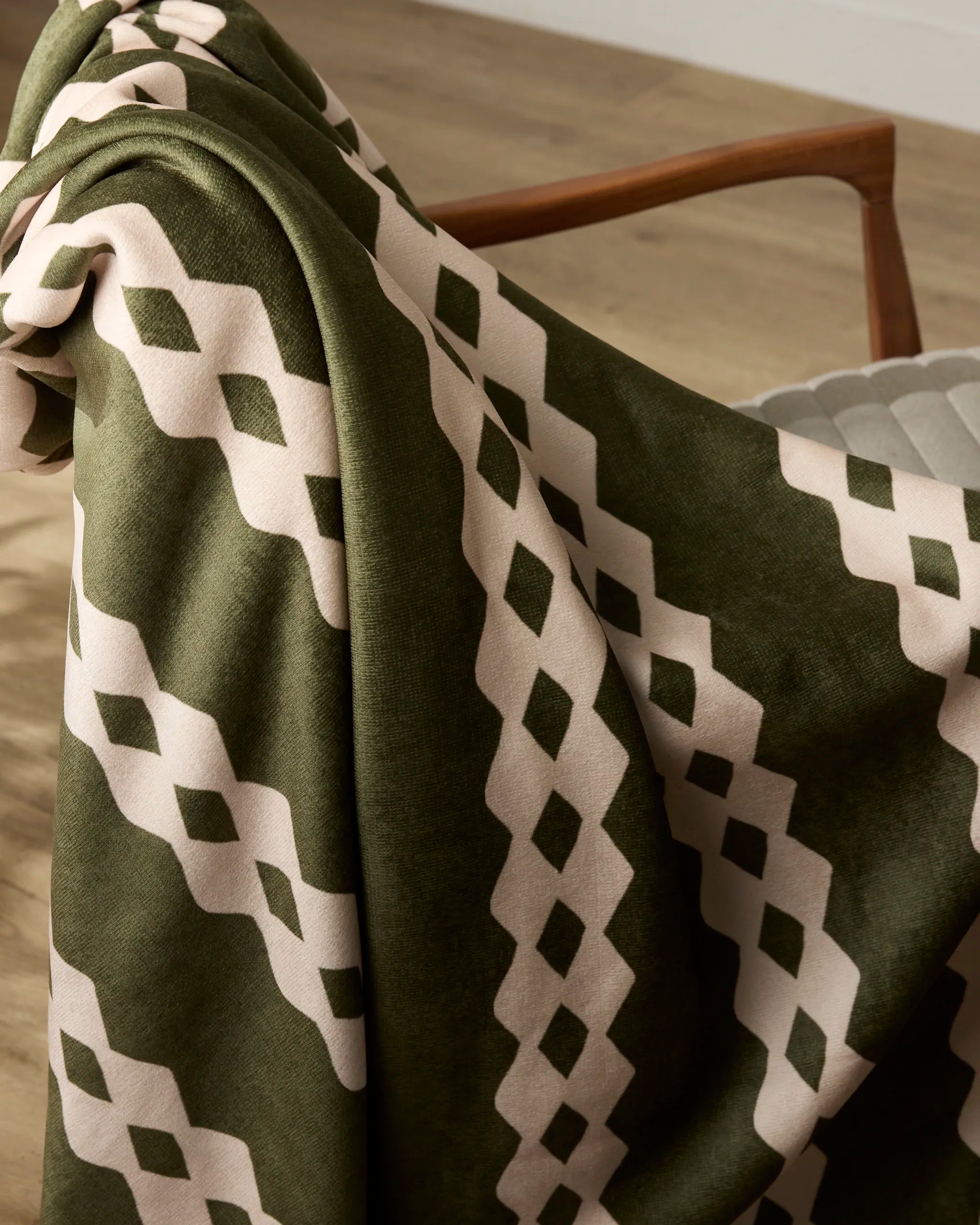 The Geometry Olive Orchard throw, featuring a green and cream geometric diamond pattern, is draped over a wooden chair on a light wood floor.