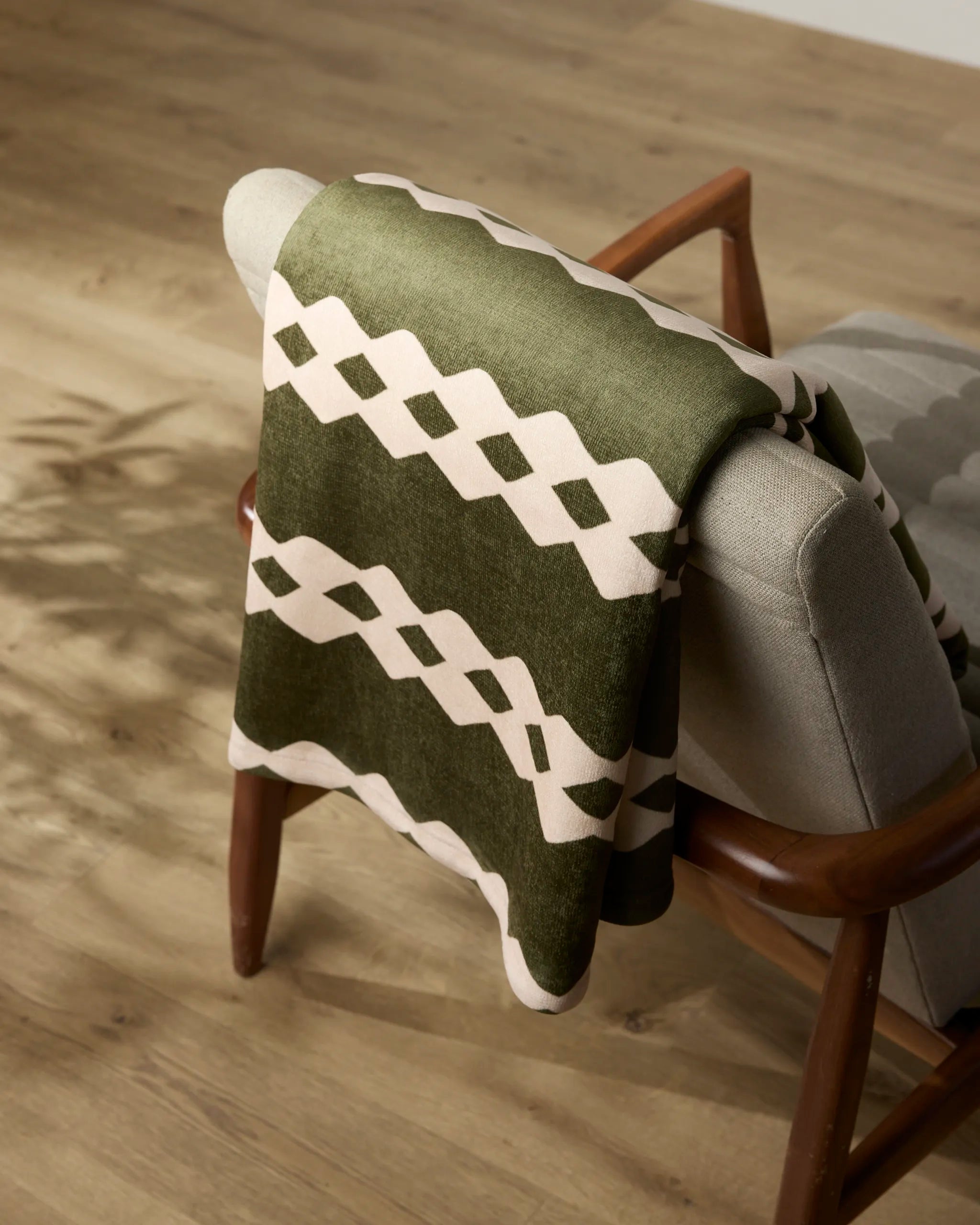 The Geometry Olive Orchard recycled blanket, featuring a green and cream geometric pattern, is draped over the back of a light gray upholstered chair with wooden arms, set on a wooden floor.