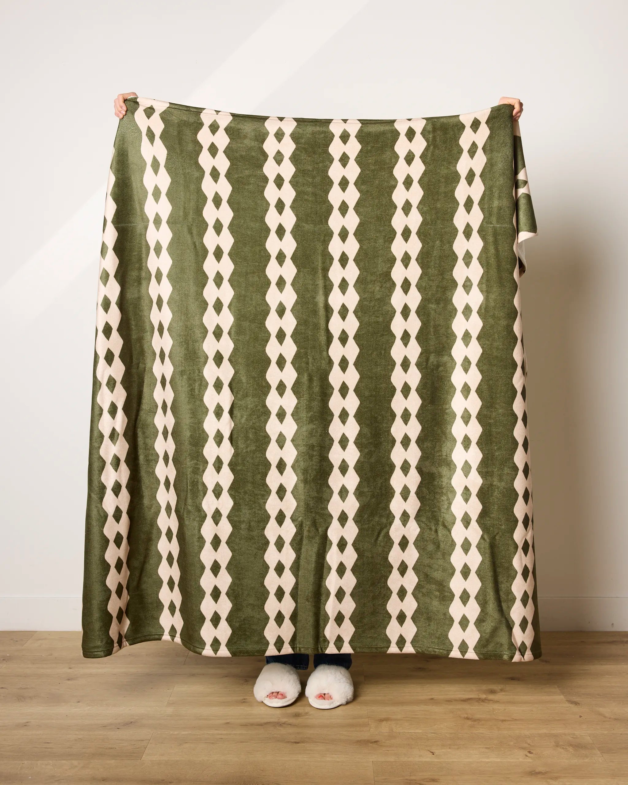 A person stands on a wooden floor, holding up Geometry's Olive Orchard blanket—an ultra-soft, recycled throw featuring olive green and cream diamond stripes. Only their hands and white-slippered feet are visible.
