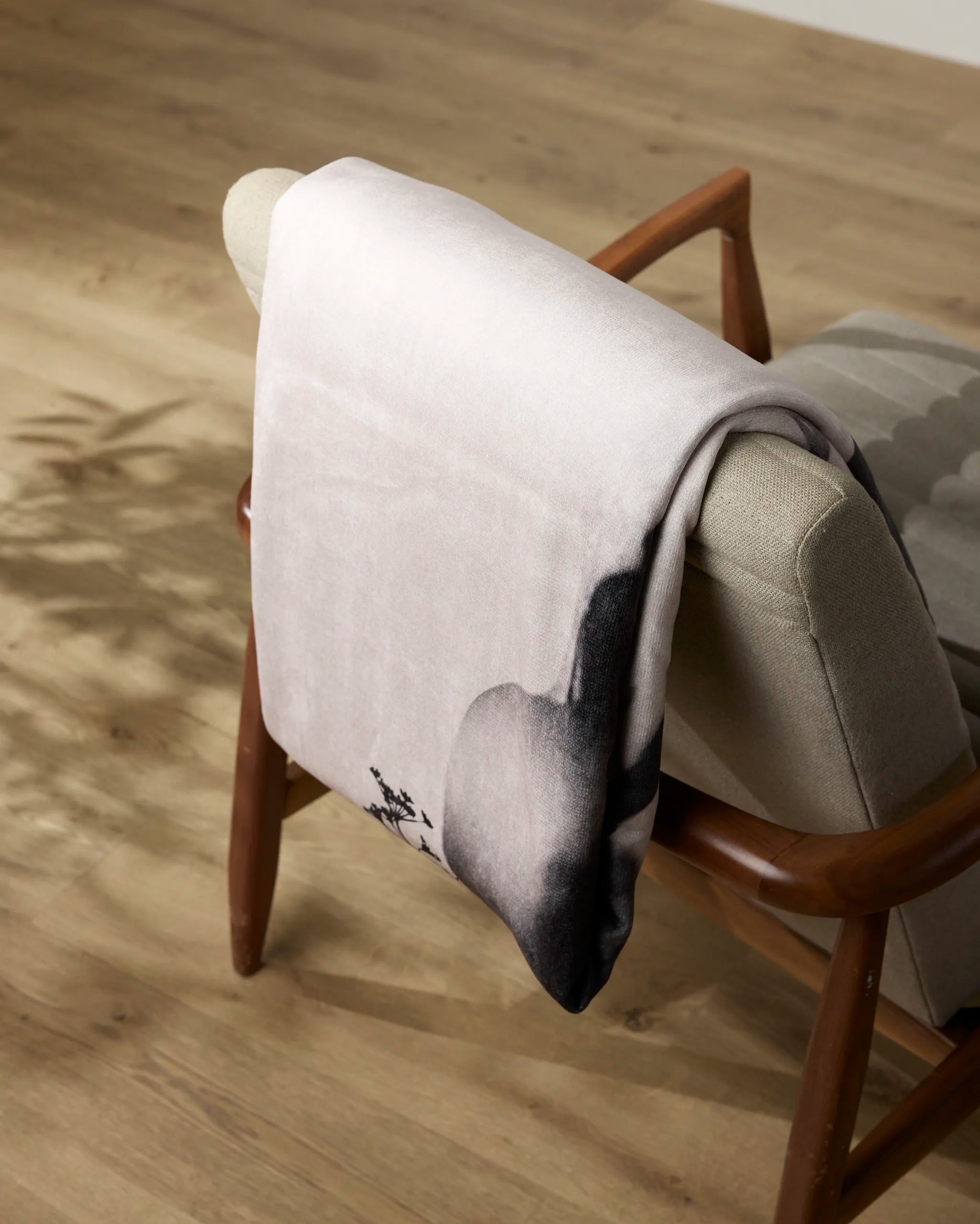 The Regal Butter Blanket by Geometry, featuring a dark abstract pattern, is draped over a cushioned armchair with wooden legs and armrests on a light wooden floor.