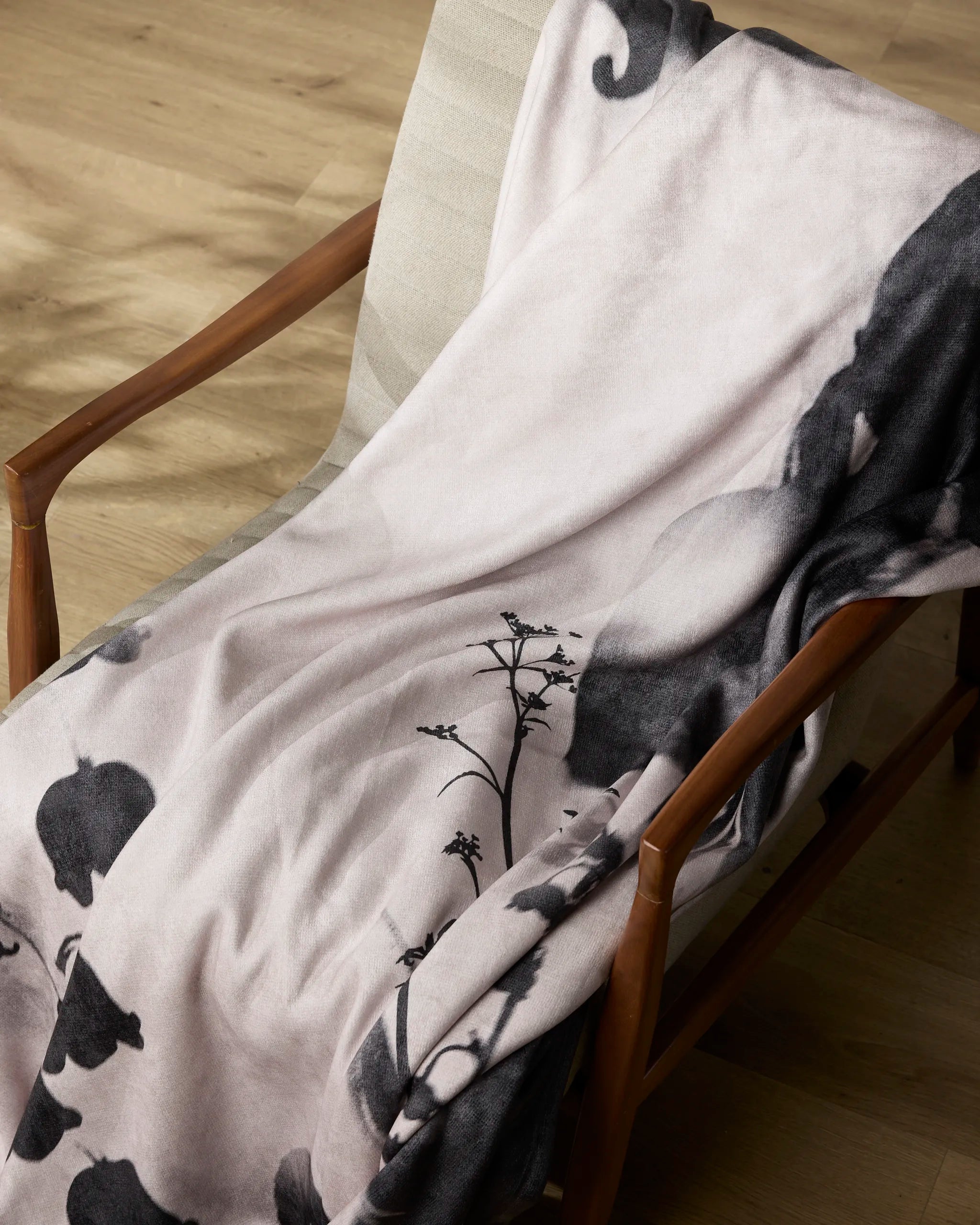 A soft Regal throw blanket by Geometry in beige and black with a floral and abstract design, draped over a wooden chair with a light cushion, brings cozy charm to any space on a light wood floor.