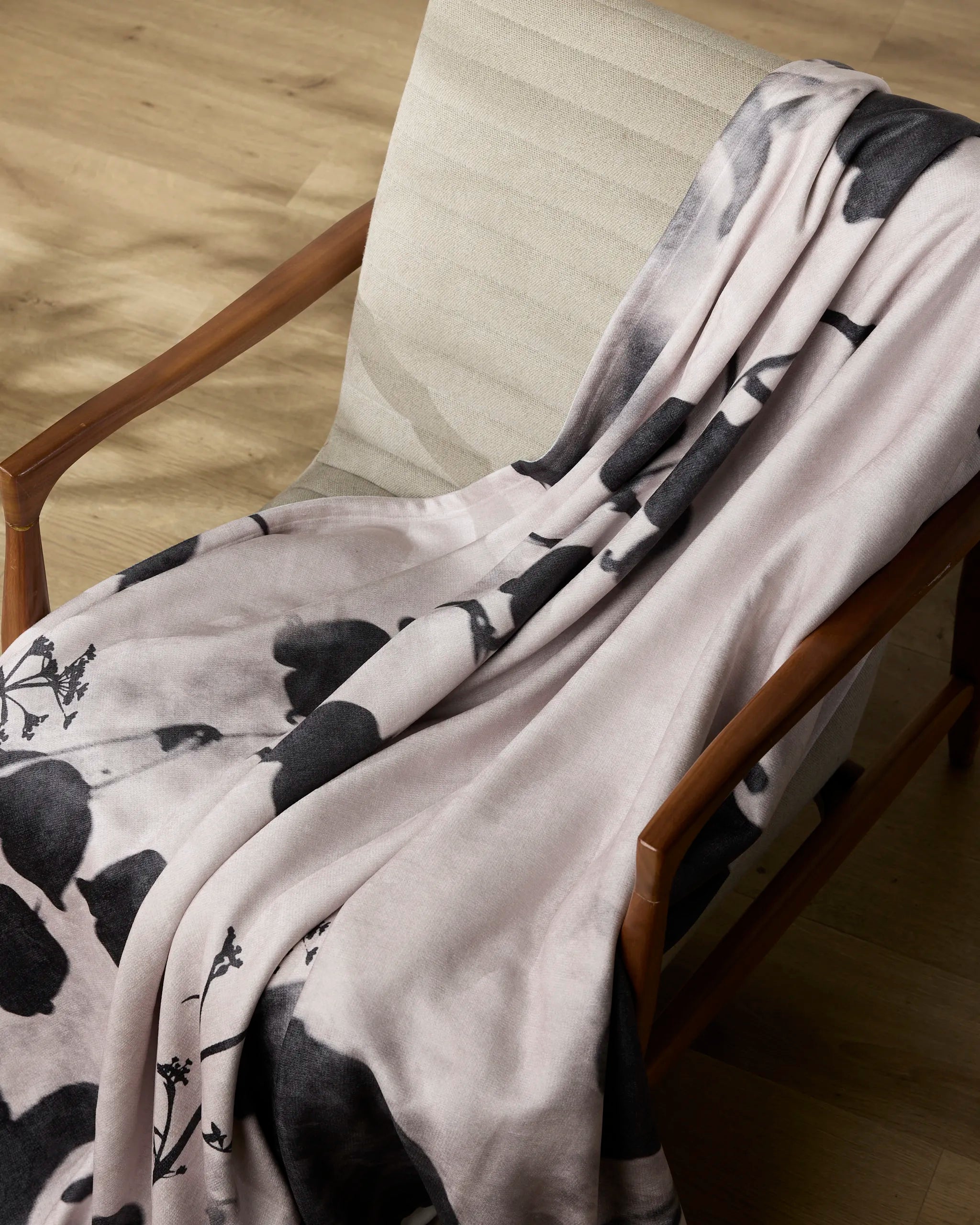 The Geometry Regal throw, featuring a soft black floral design, is draped over a cushioned wooden chair on a wooden floor.