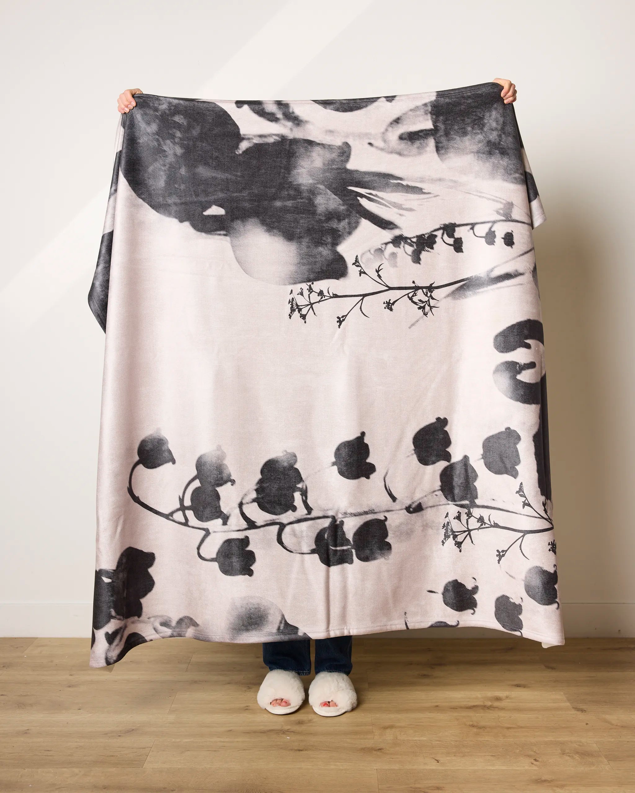 A person holds up the Geometry Regal blanket, featuring an ultra-soft black and white floral pattern. Standing on a wooden floor against a light-colored wall, only their hands, slippers, and lower legs are visible.