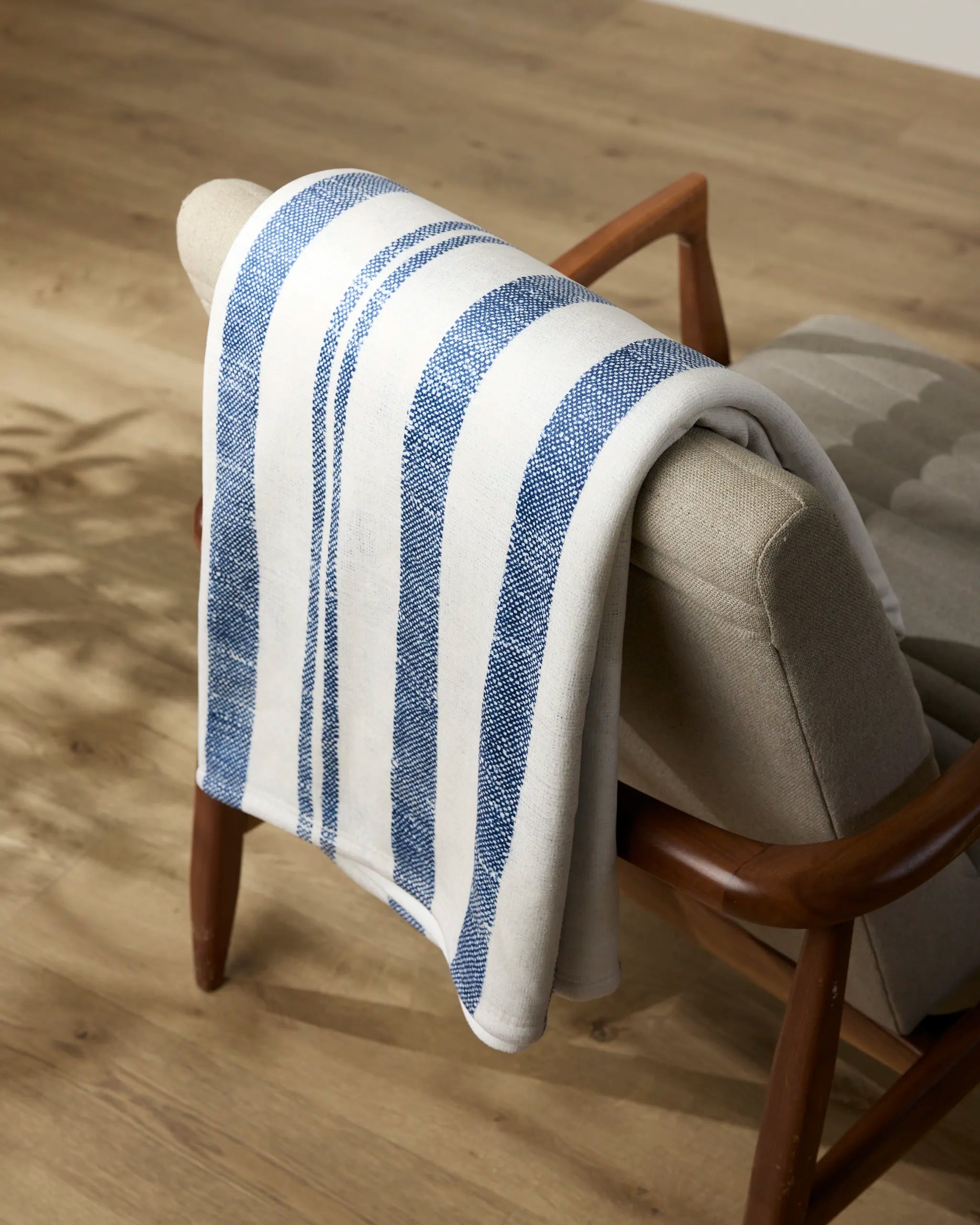 A silky soft Rhodes Butter Blanket by Geometry, featuring blue stripes, is draped over a light beige upholstered chair with wooden arms on a light wood floor.