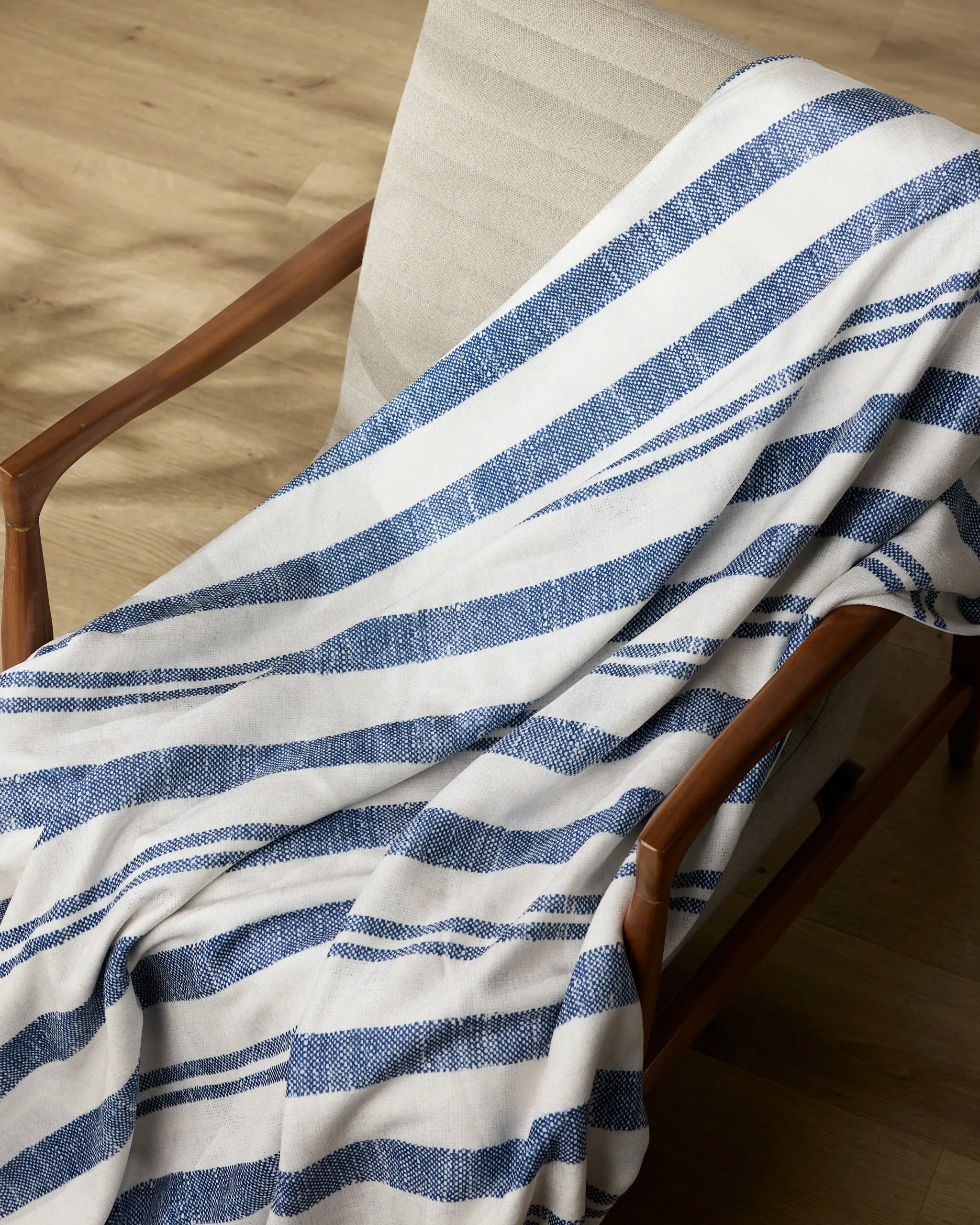 The Rhodes blanket by Geometry, featuring blue and white stripes and made from recycled materials, adds silky softness as it drapes over a wooden chair with a beige cushion on a light wood floor.
