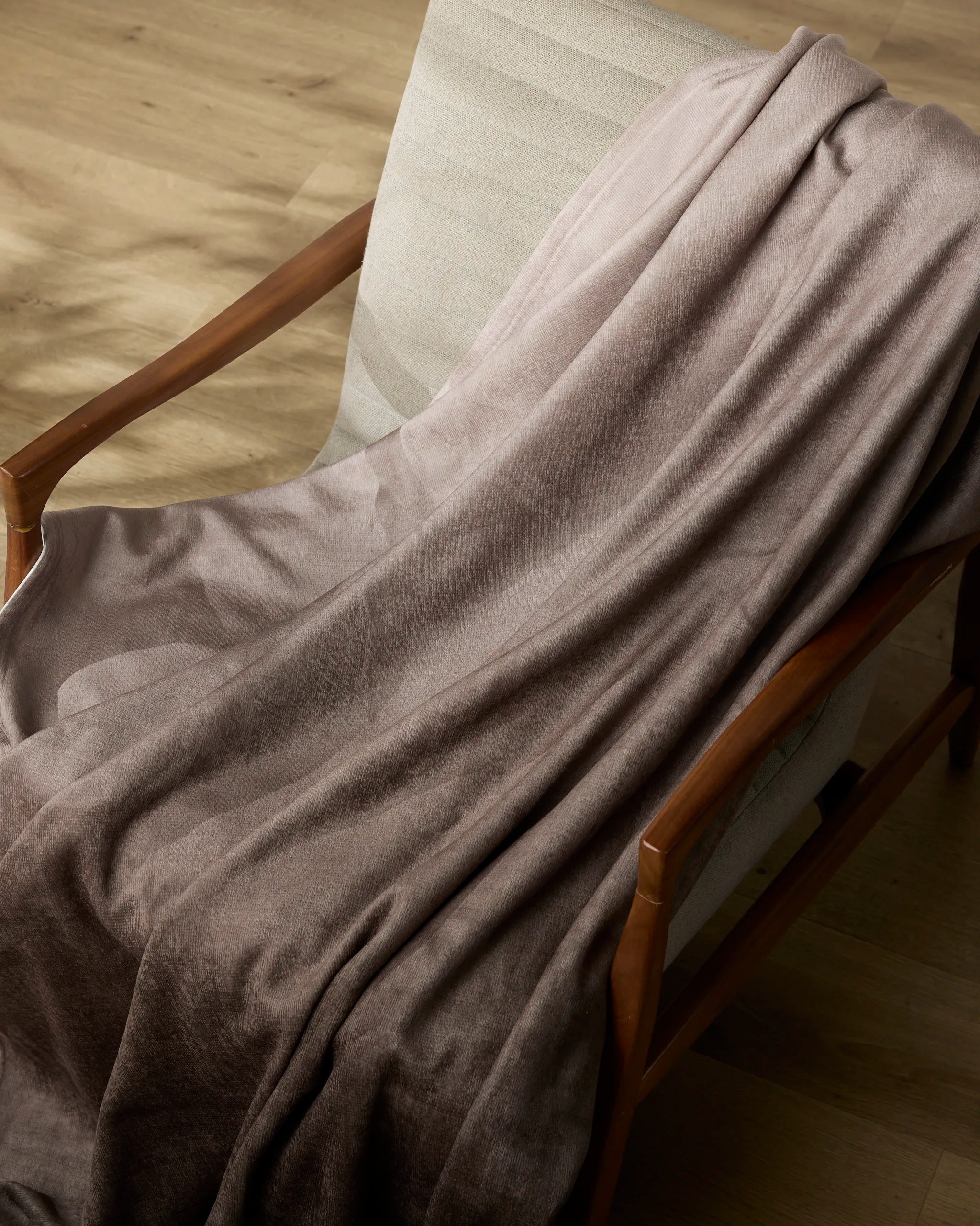A Ronan blanket by Geometry, soft and light brown, is draped over a cushioned wooden armchair placed on a light wooden floor.