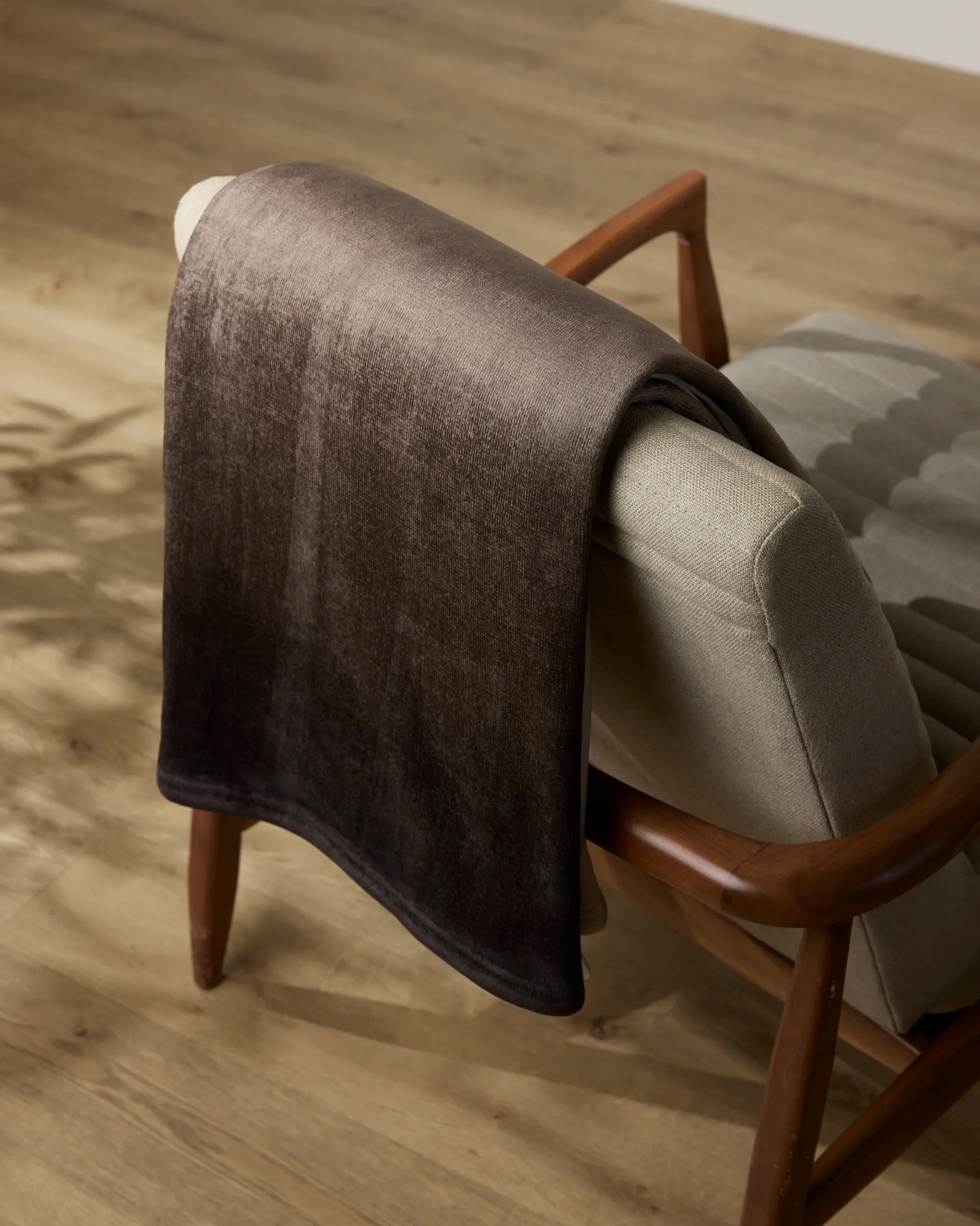 A soft, dark brown Ronan throw blanket from Geometry is draped over the back of a light gray upholstered chair with wooden arms, set on a light wood floor in a minimal, cozy room.