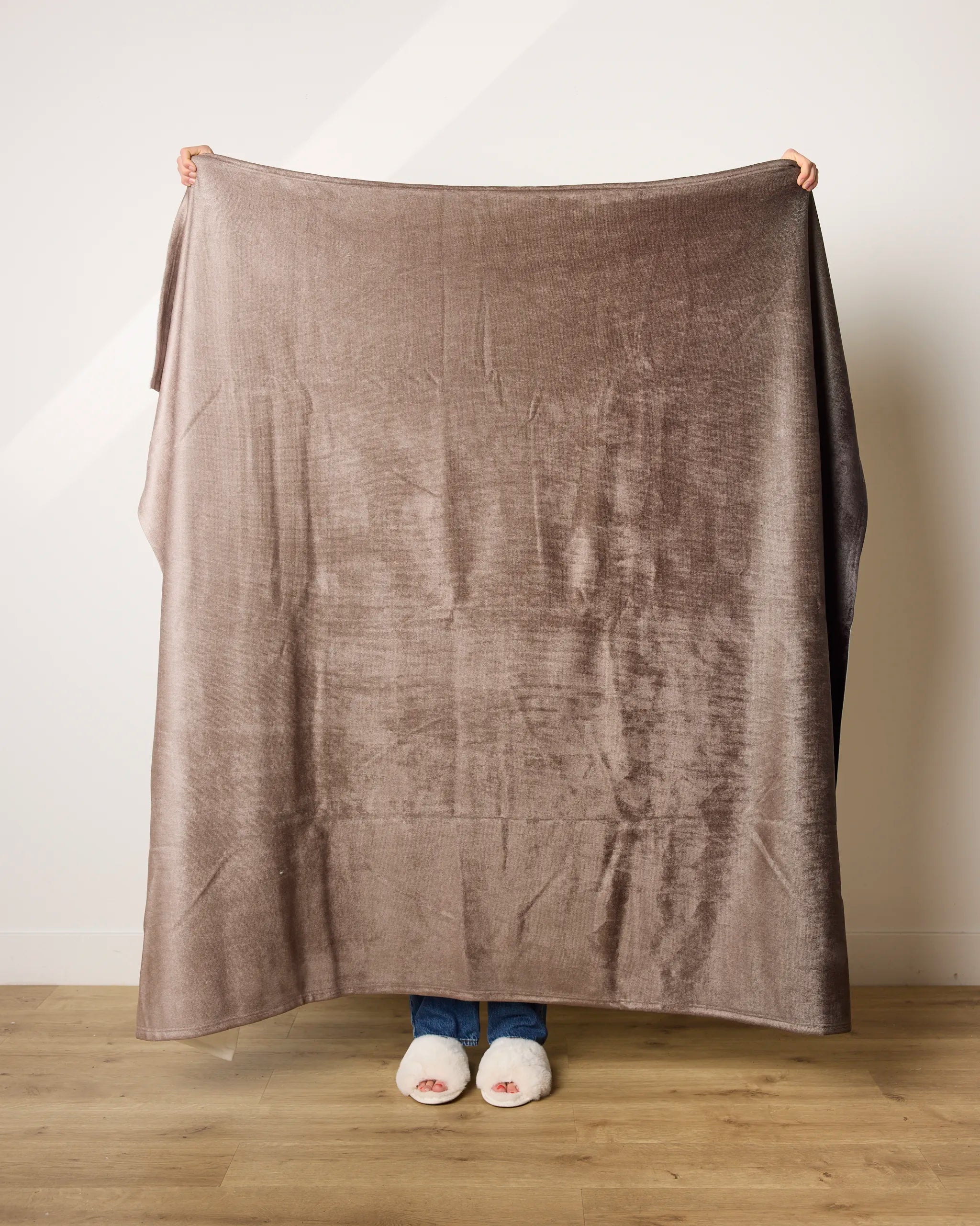 A person stands on a wooden floor holding up the ultra-soft Ronan blanket by Geometry, made from recycled materials; only their slippered feet and fingertips are visible behind it.