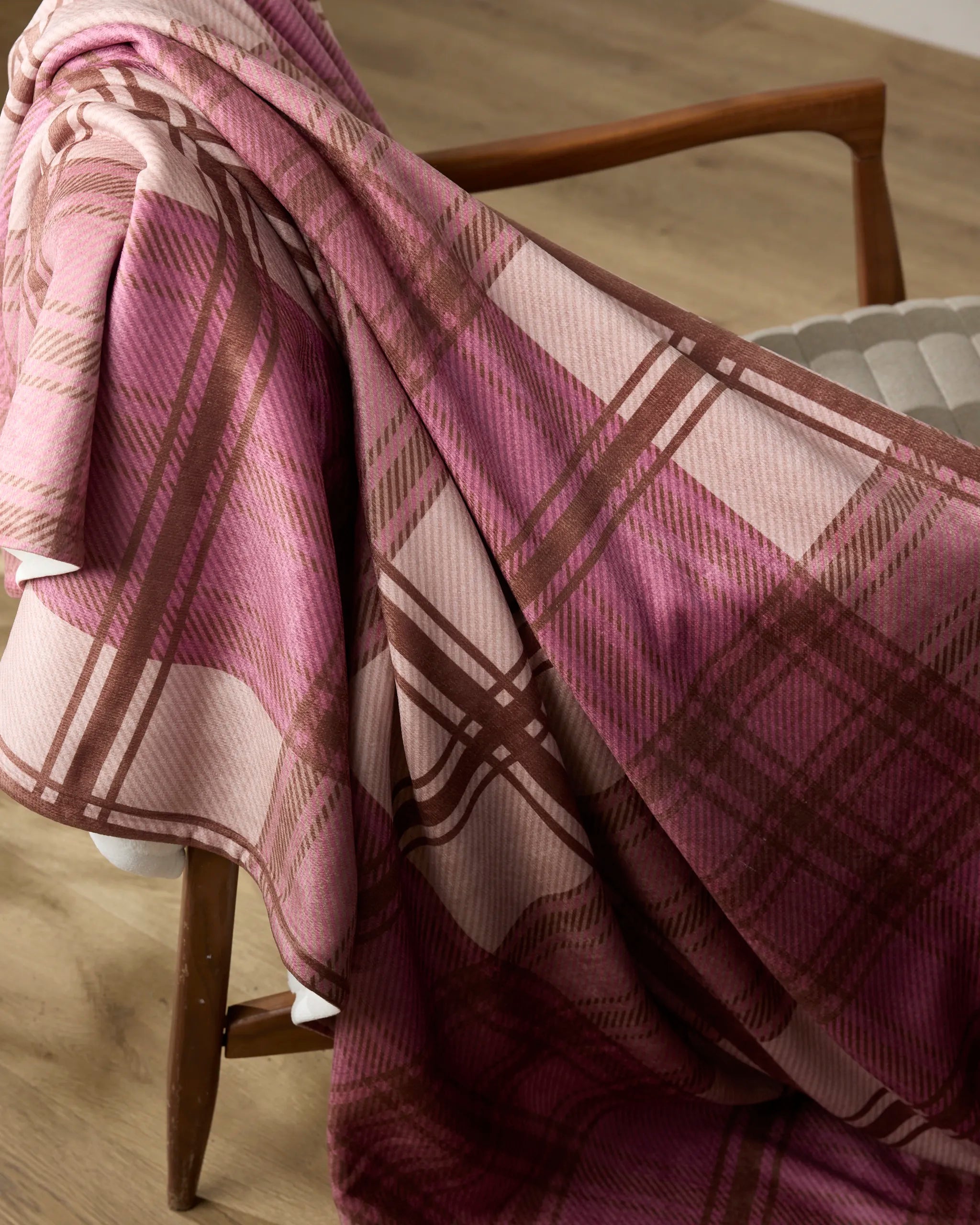 The Geometry Rosewood Tartan throw blanket is draped over the arm of a cushioned wooden chair on a light wood floor.