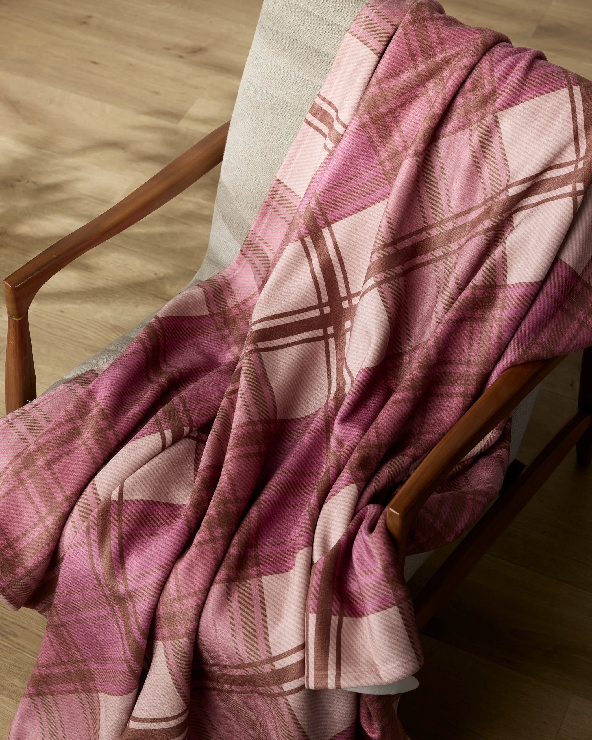 The Geometry Rosewood Tartan recycled throw blanket, featuring a pink plaid design with brown accents, is draped over a light-cushioned wooden chair on a light wood floor—perfect for cozy moments.