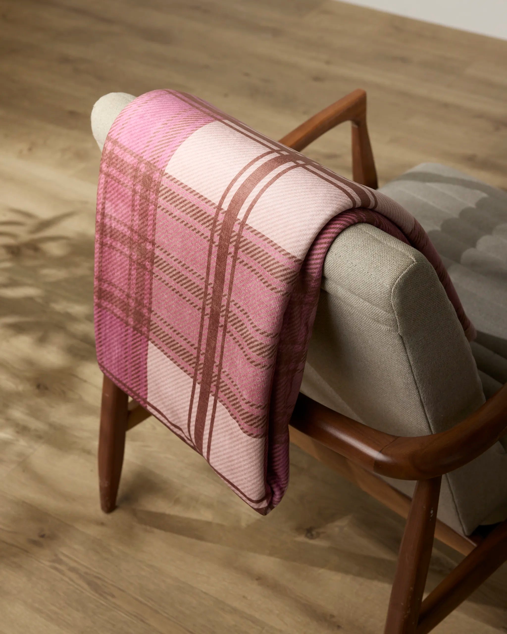 The Geometry Rosewood Tartan throw blanket is draped over the back of a light gray upholstered wooden chair on a wooden floor.
