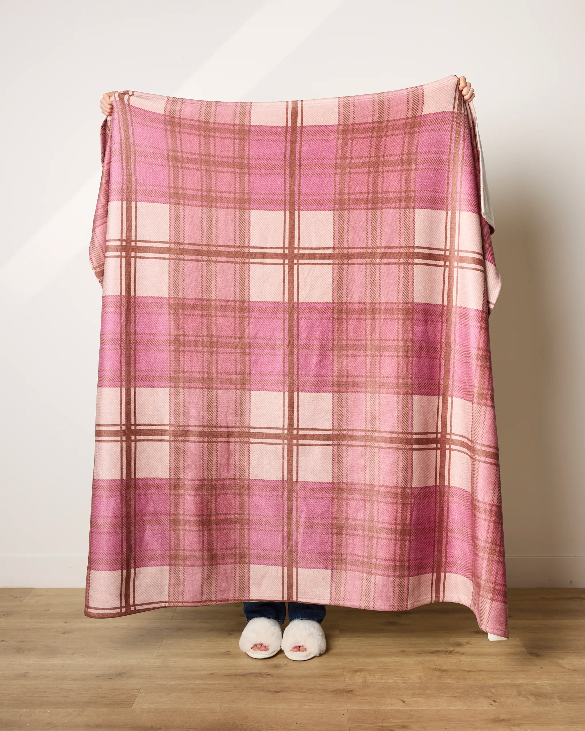 A person stands indoors wrapped in the ultra-soft Geometry Rosewood Tartan blanket in pink and white plaid, covering themselves except for their hands and feet in white slippers, against a plain wall and wooden floor.
