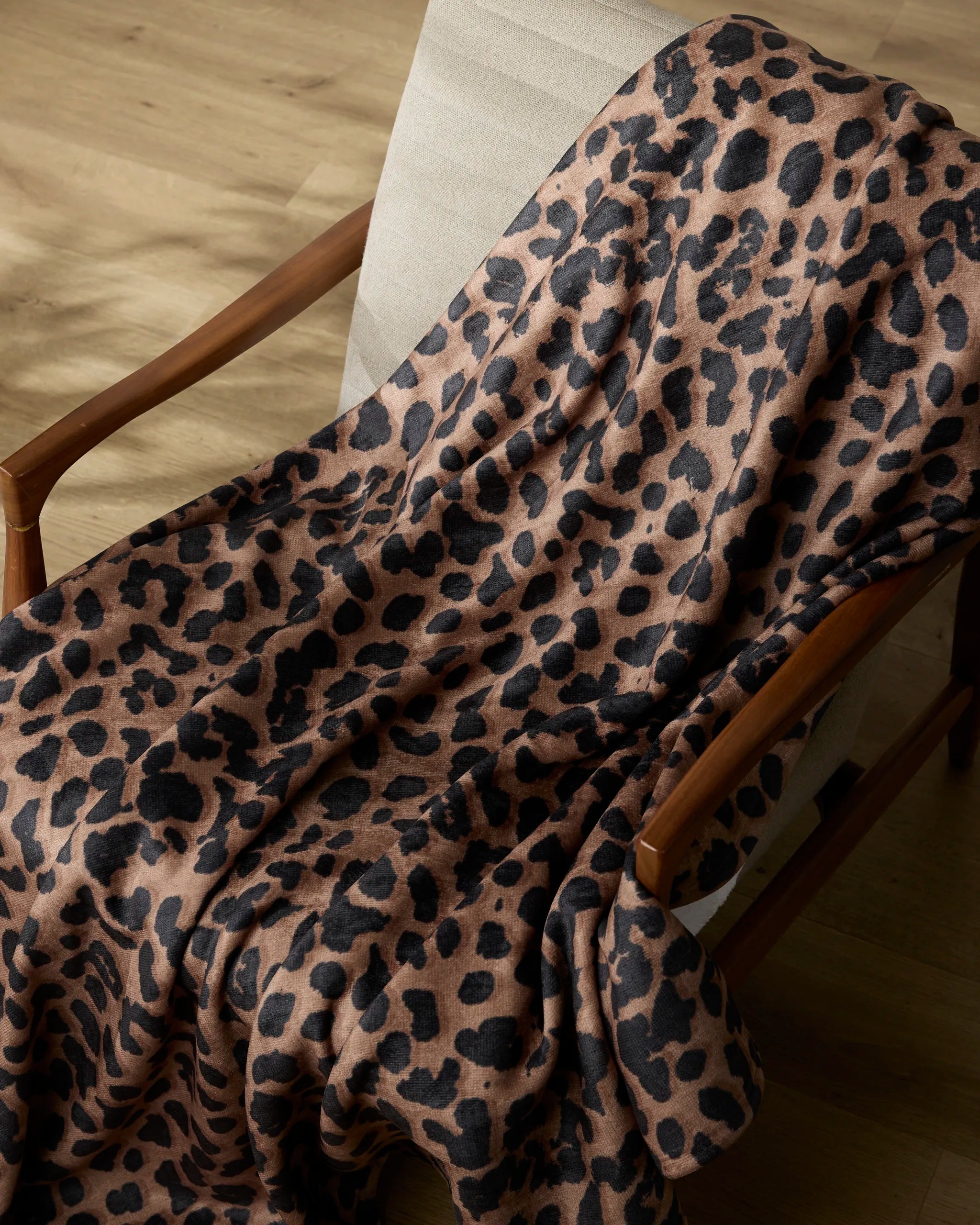 The Geometry Savannah throw blanket, featuring a black leopard print, is draped over a light beige cushioned wooden chair on a wooden floor, bringing cozy style to any space.