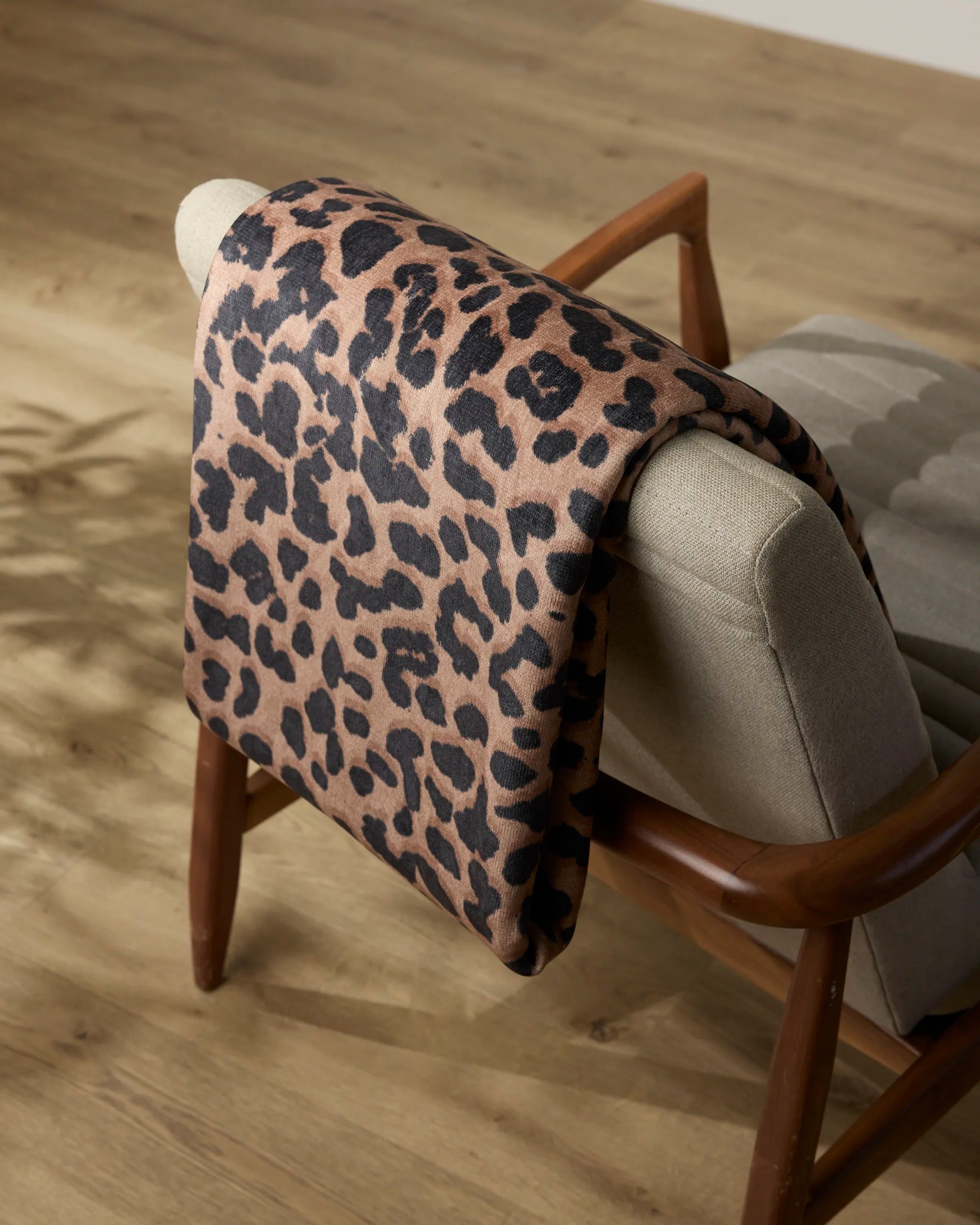 The Geometry Savannah throw blanket, featuring a brown base with black leopard spots, is draped over the back of a light gray upholstered wooden chair on a light wood floor.