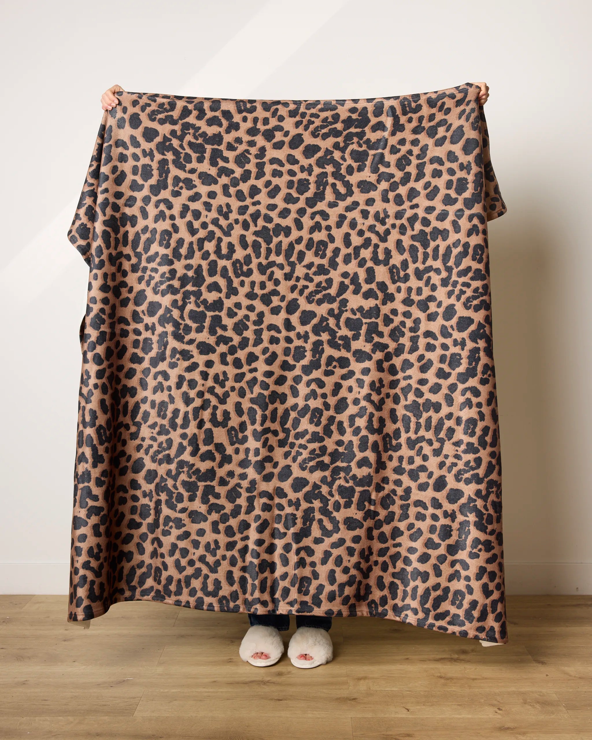 A person stands on a wooden floor, mostly hidden behind the Savannah blanket by Geometry, made from recycled materials in brown and black tones. Only their hands and feet are visible against a plain white wall.