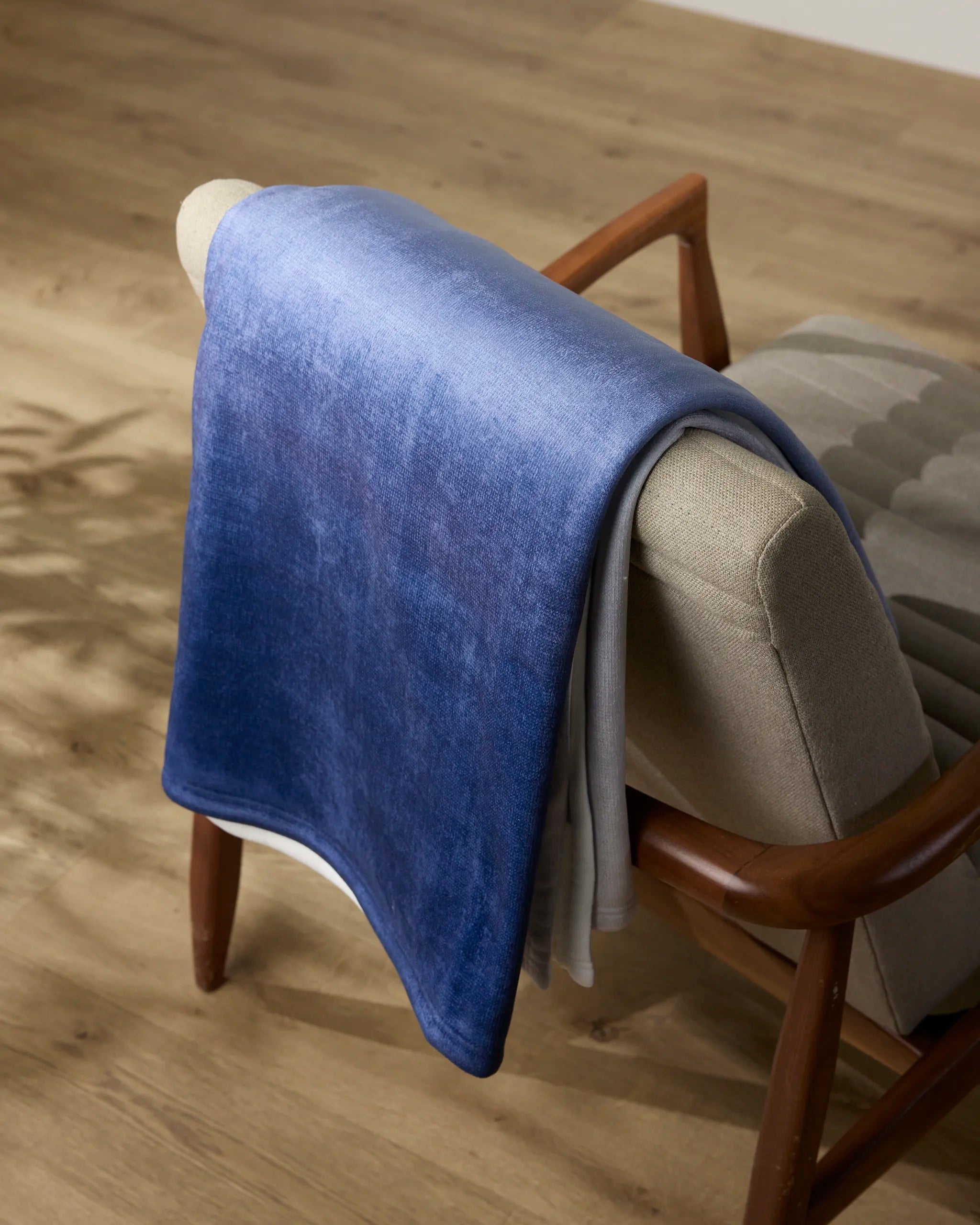 The Geometry Soft Skies throw blanket in a blue gradient is draped over the backrest of a modern beige armchair with wooden arms, situated on a light wooden floor.