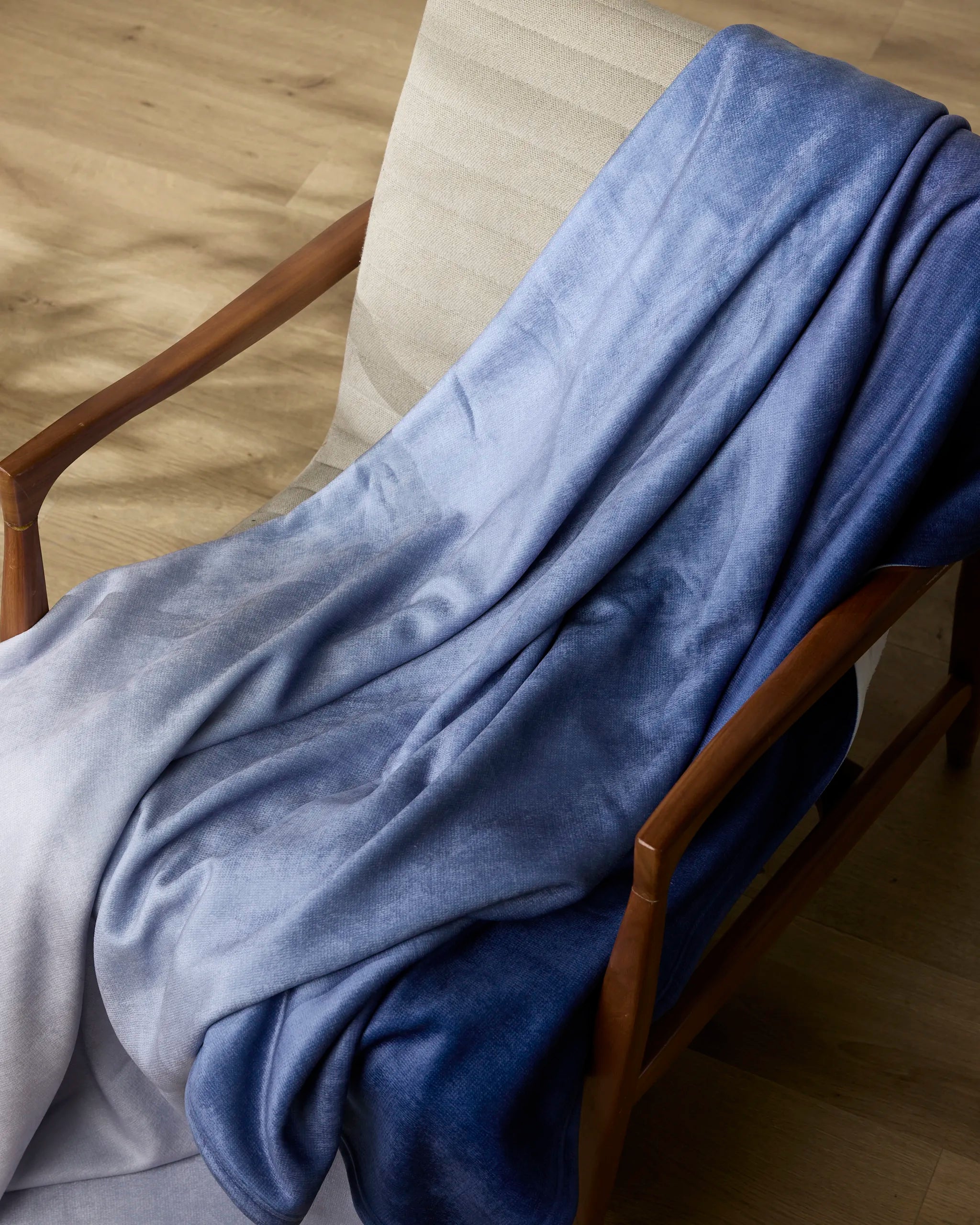 The Geometry “Soft Skies” throw blanket in gradient blue is draped over a light cushioned chair with wooden armrests, set on a wooden floor.