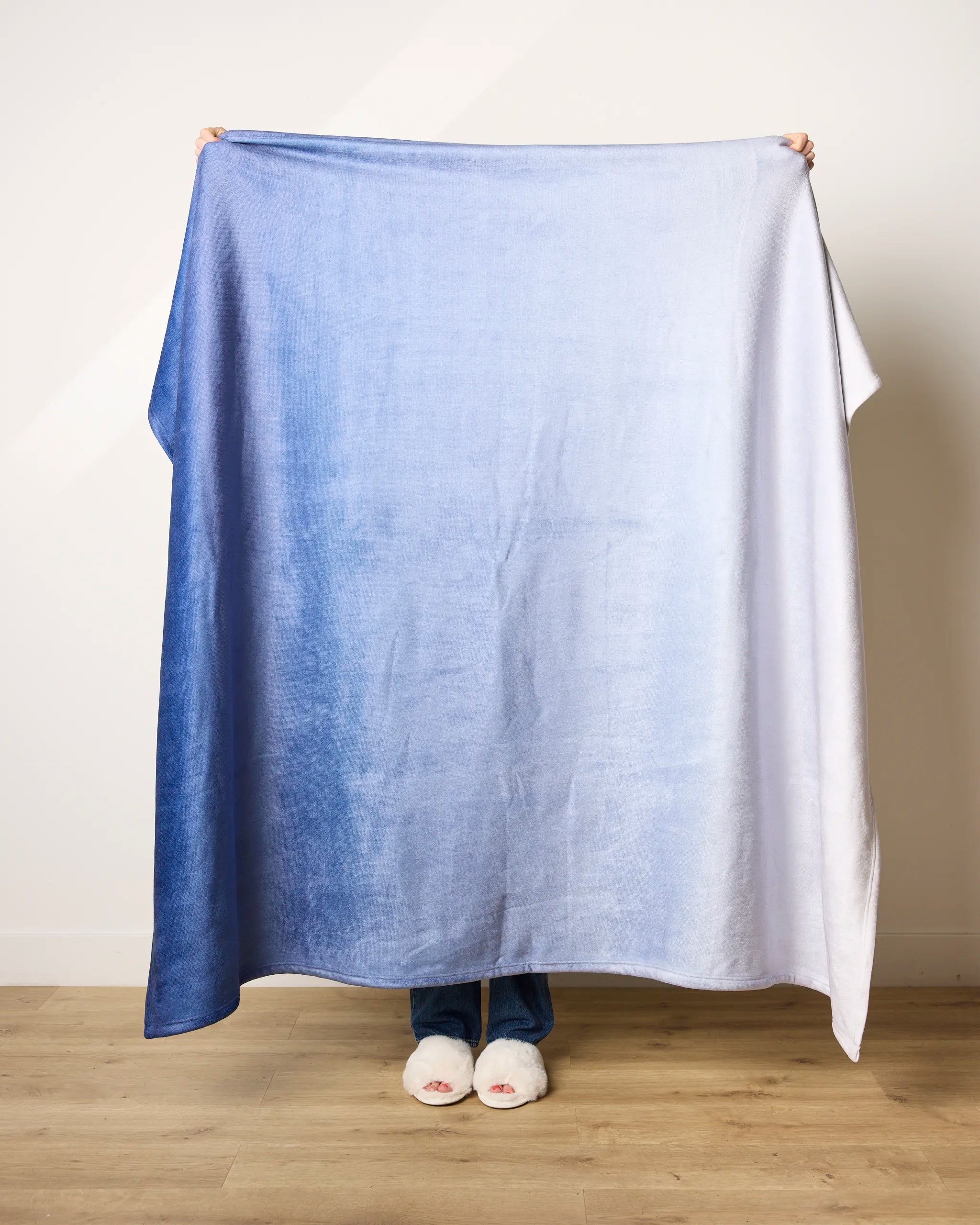 A person holds up the Geometry Soft Skies blanket, featuring an ultra-soft blue-to-white gradient, covering their body except for hands, feet in slippers, and part of jeans, with a light wall and wooden floor in the background.