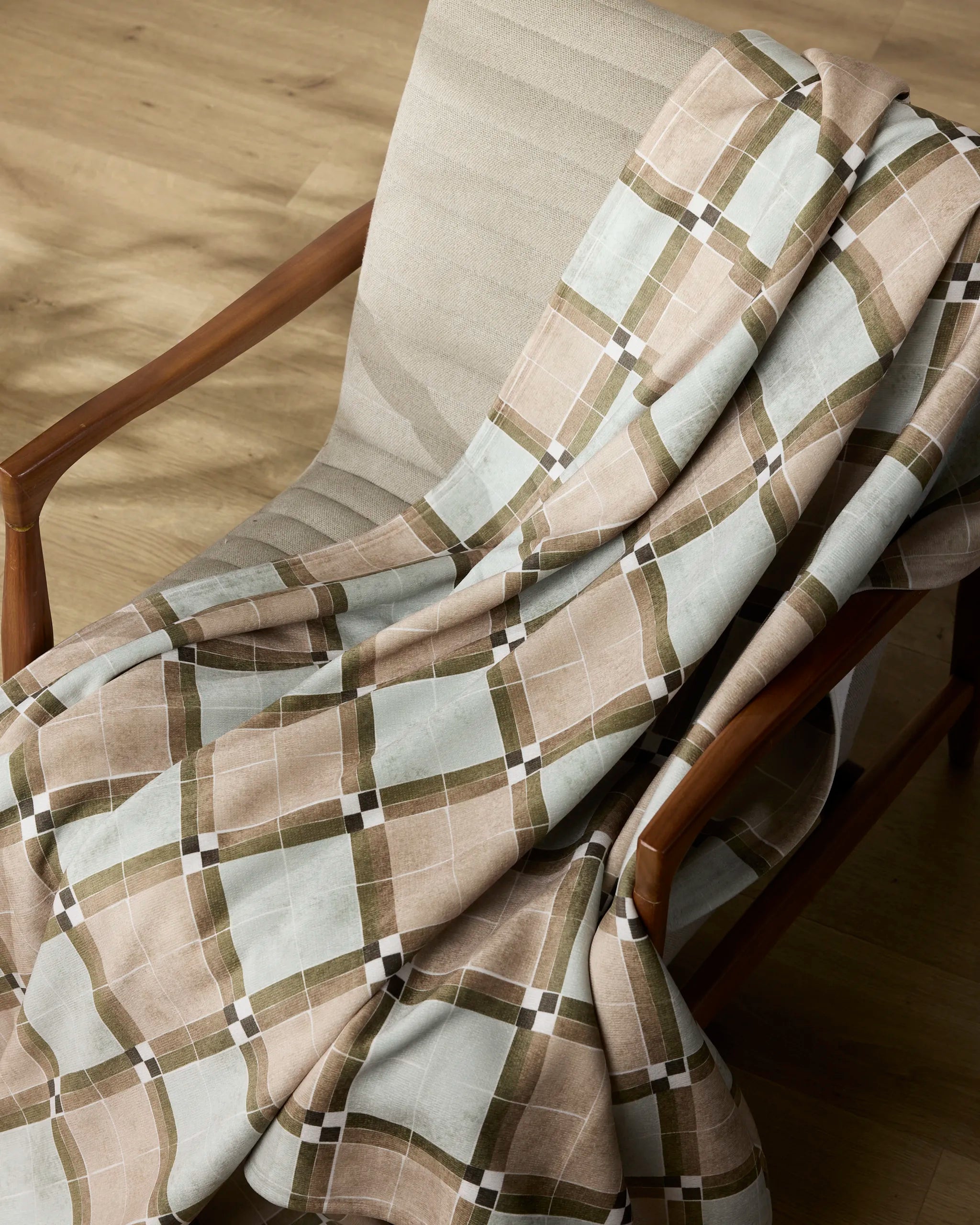 The Sunday Morning Butter Blanket by Geometry, crafted from recycled blankets in beige and olive green, is draped over a light-upholstered chair with wood arms, resting on a light wood floor.