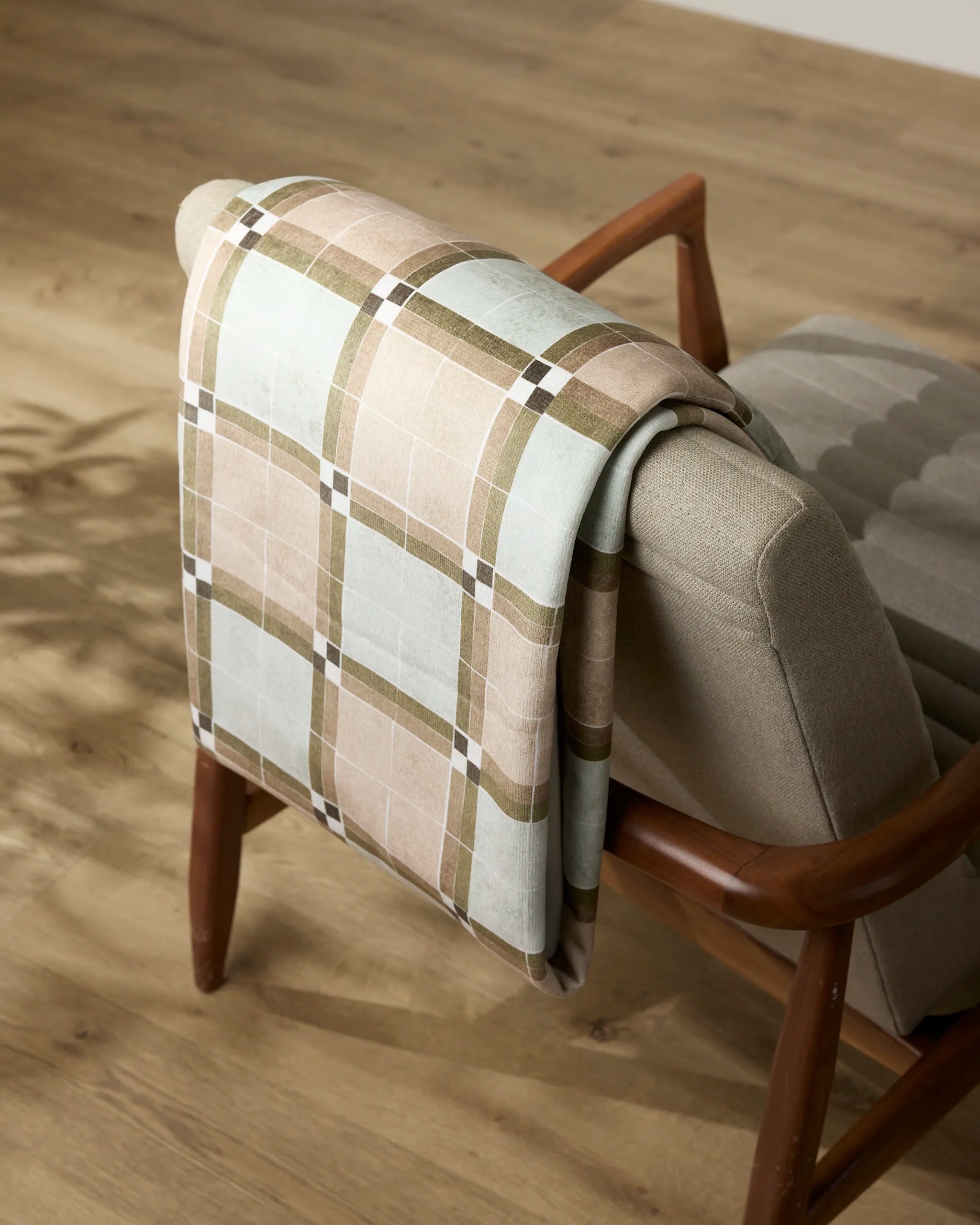 A Sunday Morning blanket by Geometry, soft and cozy, is draped over the back of a mid-century modern armchair with wooden arms, set on a light wood floor.
