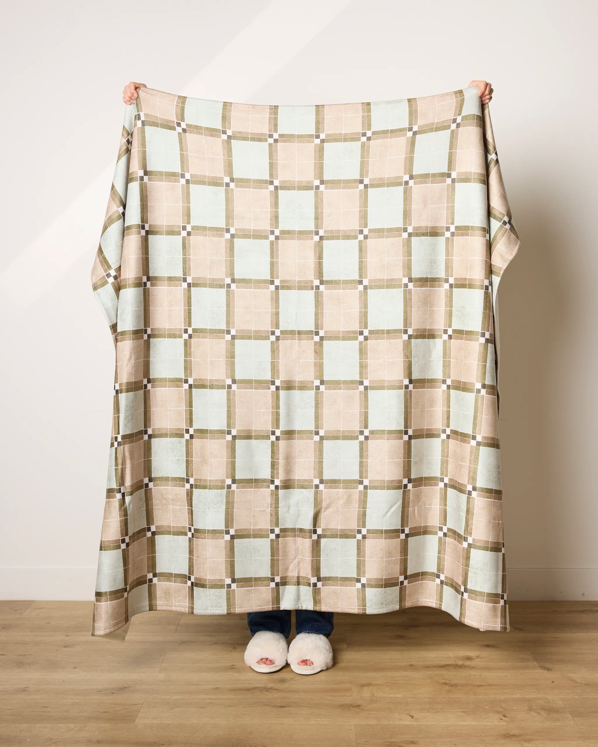 A person stands indoors holding up the Sunday Morning blanket by Geometry, featuring beige, brown, and light blue squares. Only their hands, jeans, and white slippers are visible. This ultra-soft blanket is made from recycled materials.