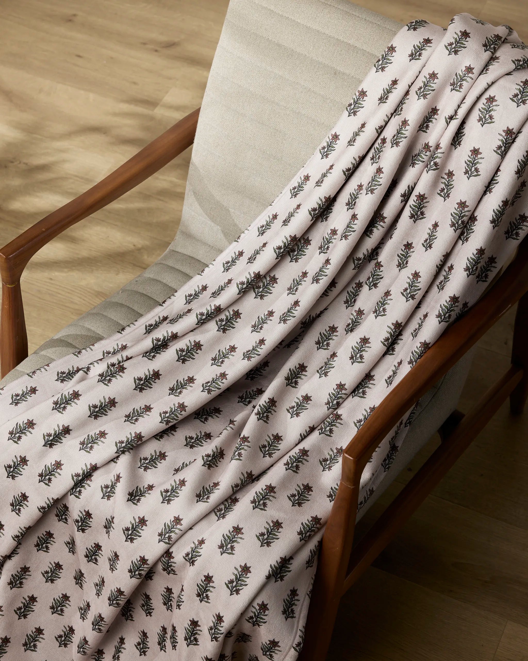 The Sweet Lilith chair by Geometry features light beige cushioning and wooden armrests, draped with a pale pink throw blanket adorned with a subtle floral pattern, all set atop a light wood floor.