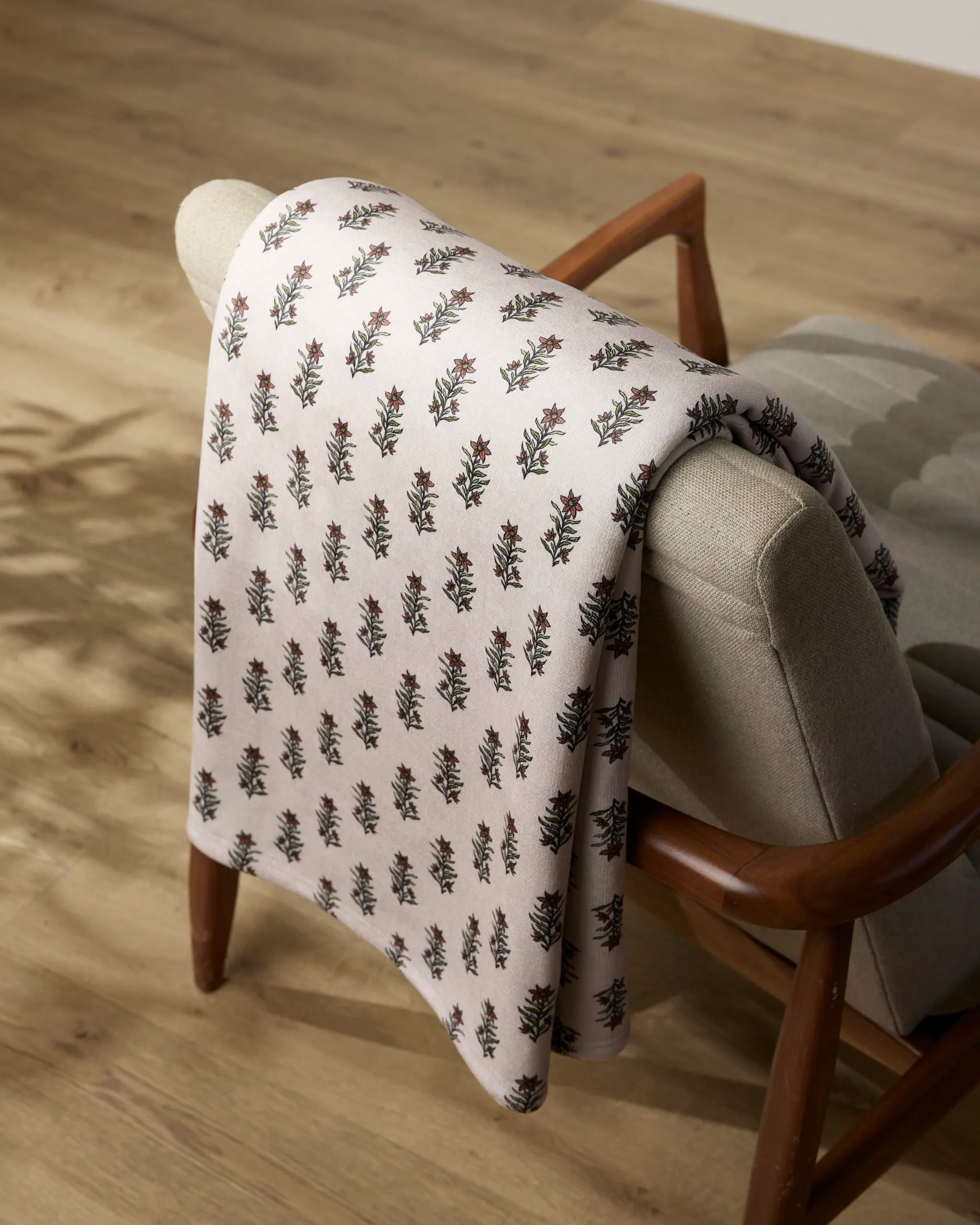 The Sweet Lilith butter blanket by Geometry, featuring a small floral pattern, is draped over the back of a light gray upholstered chair with wooden arms on a light wood floor.