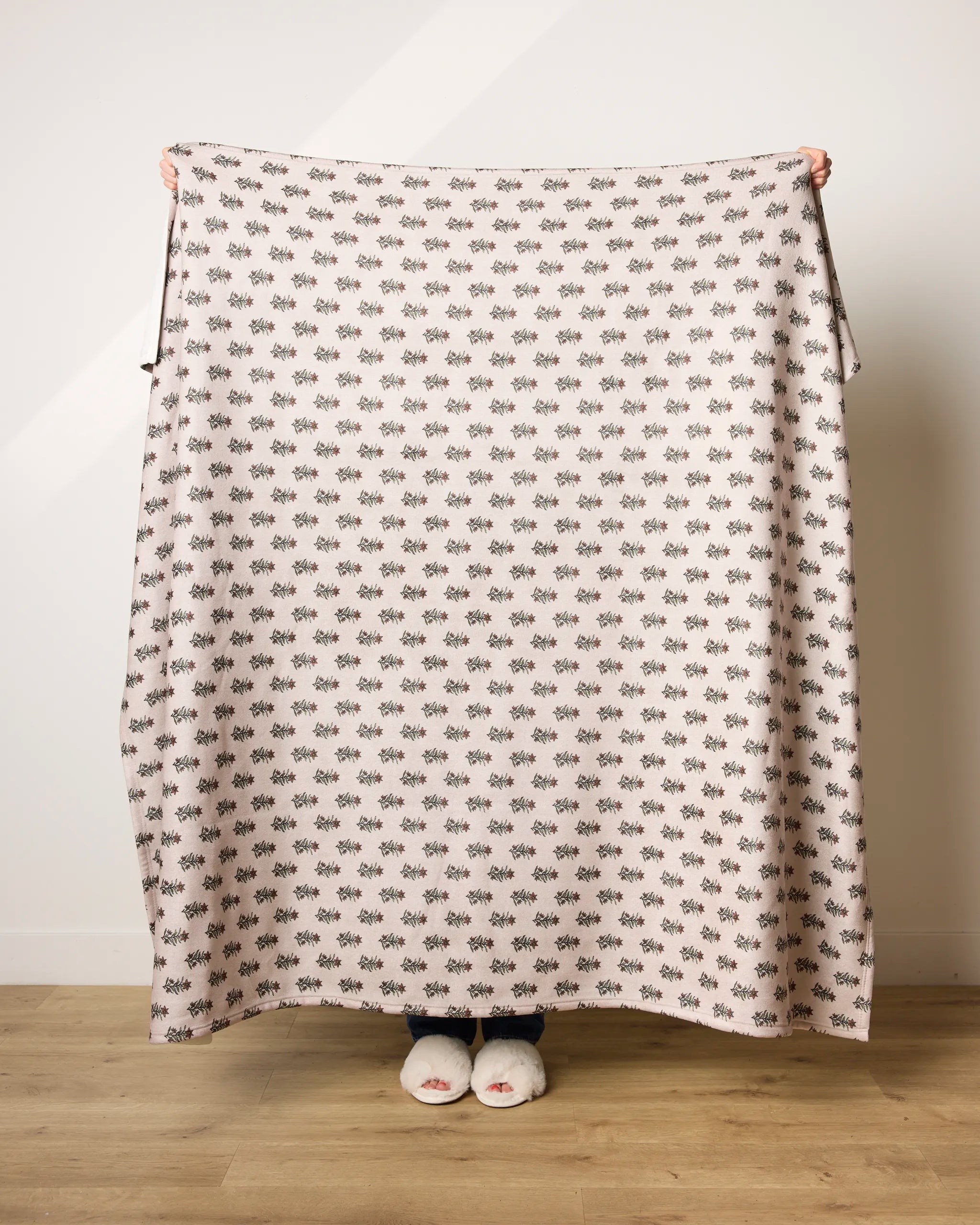 A person stands on a wooden floor, holding up Geometry's Sweet Lilith ultra-soft blanket with small dark floral motifs—only their hands and slippered feet are visible.