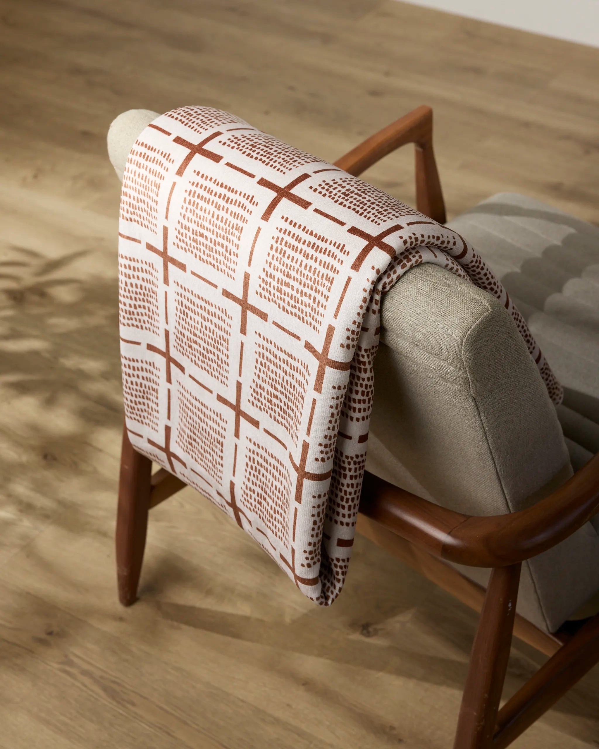 The Theo throw blanket by Geometry, featuring a beige and brown geometric pattern, is draped over a light gray upholstered wooden chair on a light wood floor.