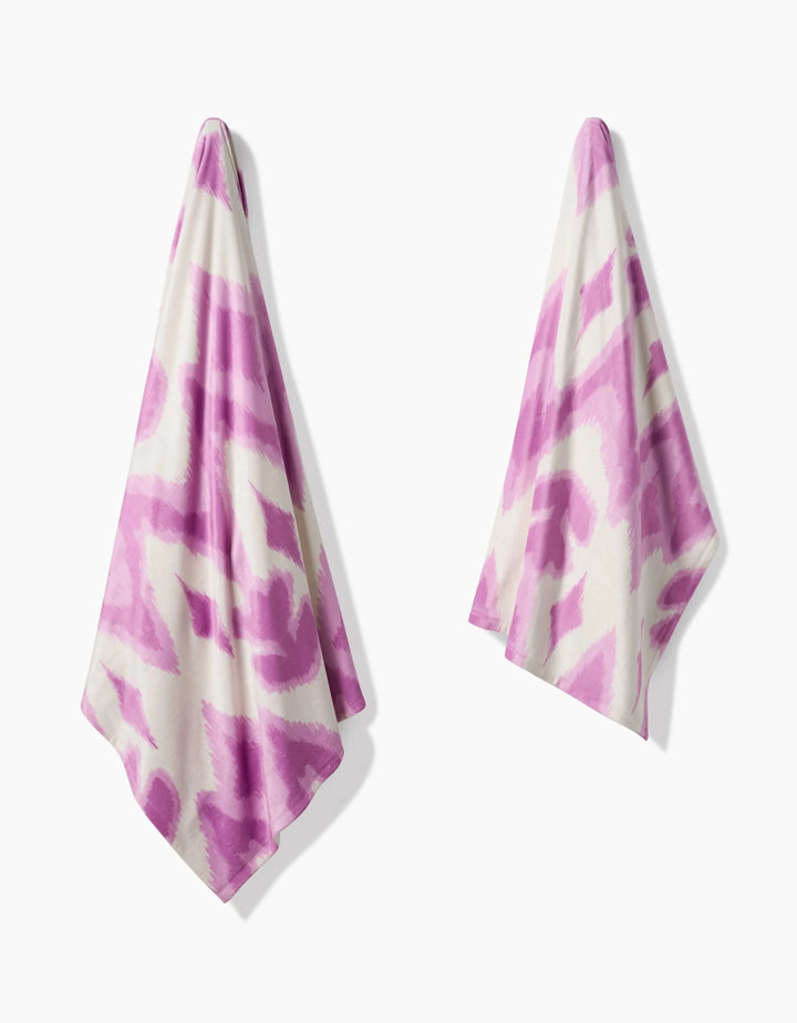 Two Tulip Scatter towels by Geometry, featuring bold abstract purple patterns, hang separately against a white background—each crafted from recycled materials for a sustainable touch.