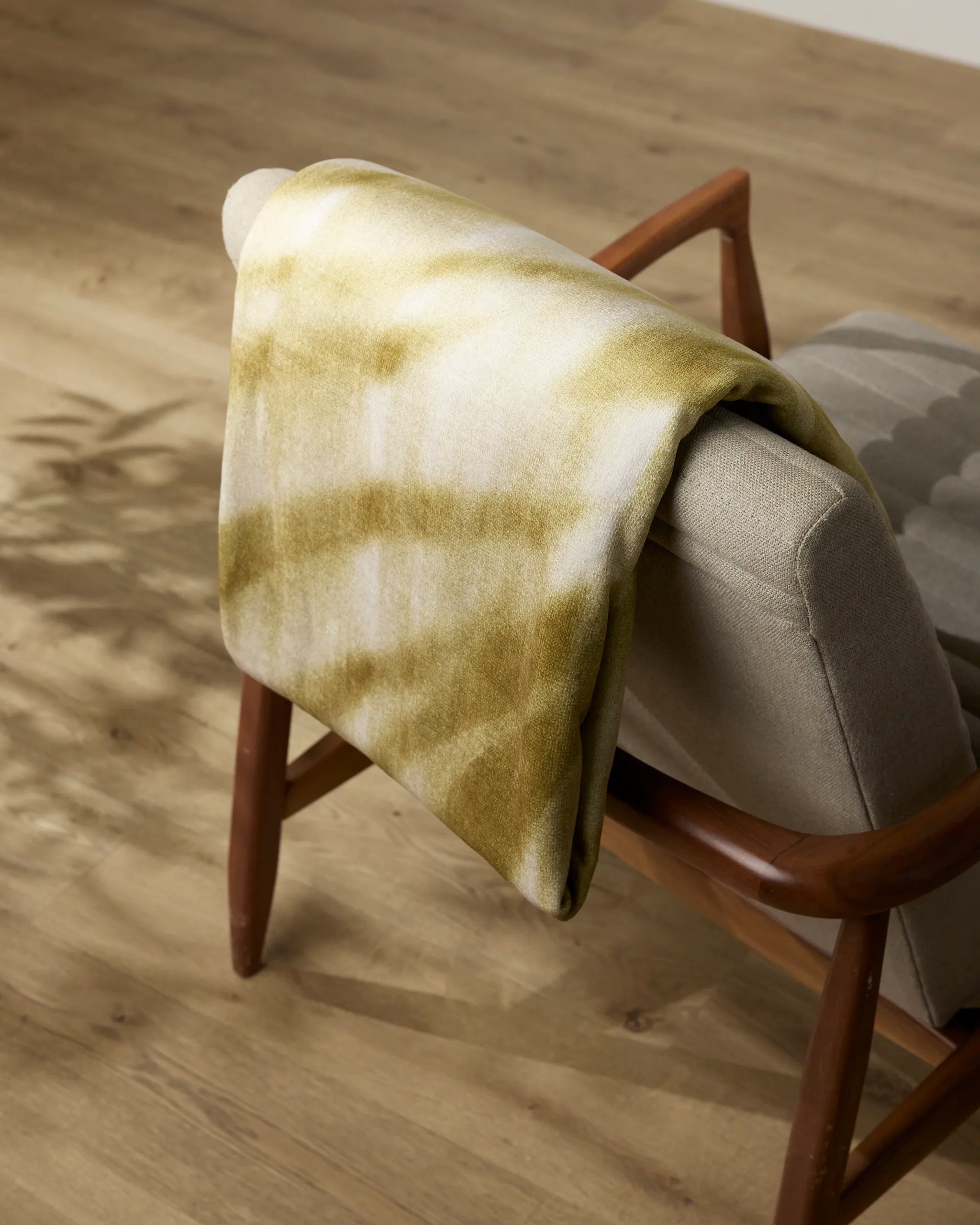 The Zain throw blanket from Geometry, featuring yellow and white tie-dye, is draped over a light gray cushioned wooden chair—perfect for cozy moments and part of our recycled blankets collection.