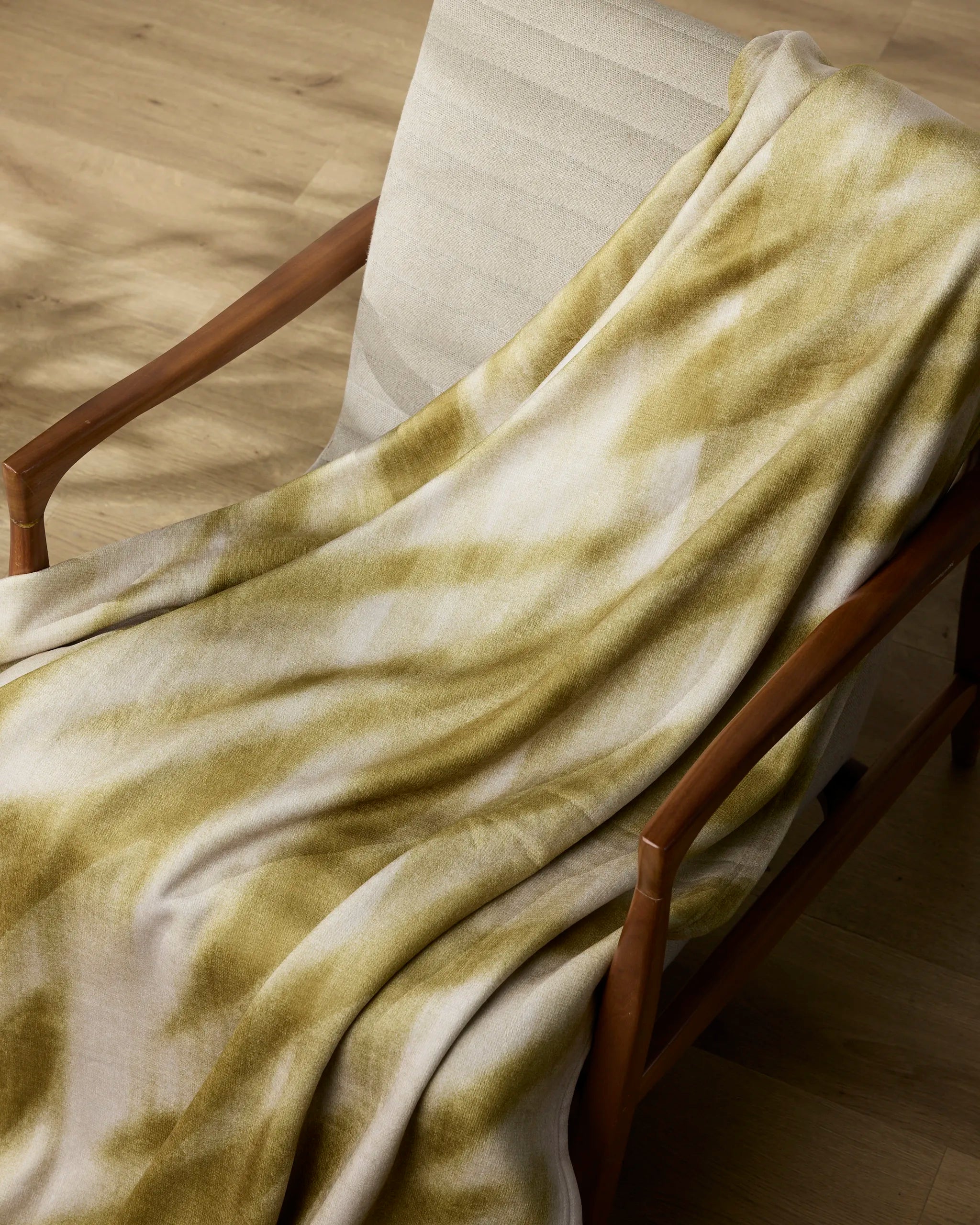 The Geometry Zain throw blanket in beige and olive green tie-dye drapes over a cushioned wooden chair on a light wood floor, with sunlight casting gentle shadows.