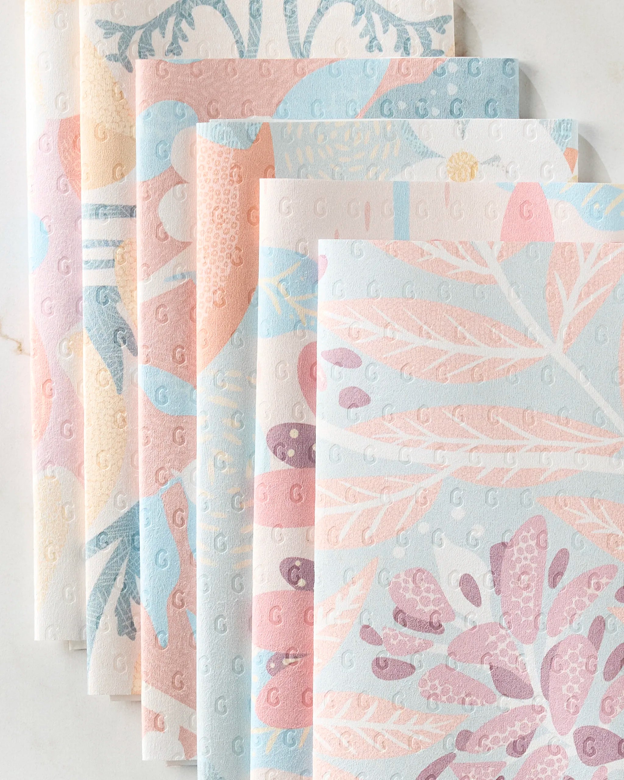 Six folded State Flowers paper napkins by Julianne Haness are displayed on a white surface, featuring pastel floral and botanical patterns in pink, blue, purple, and cream with embossed dots and subtle decorative designs.