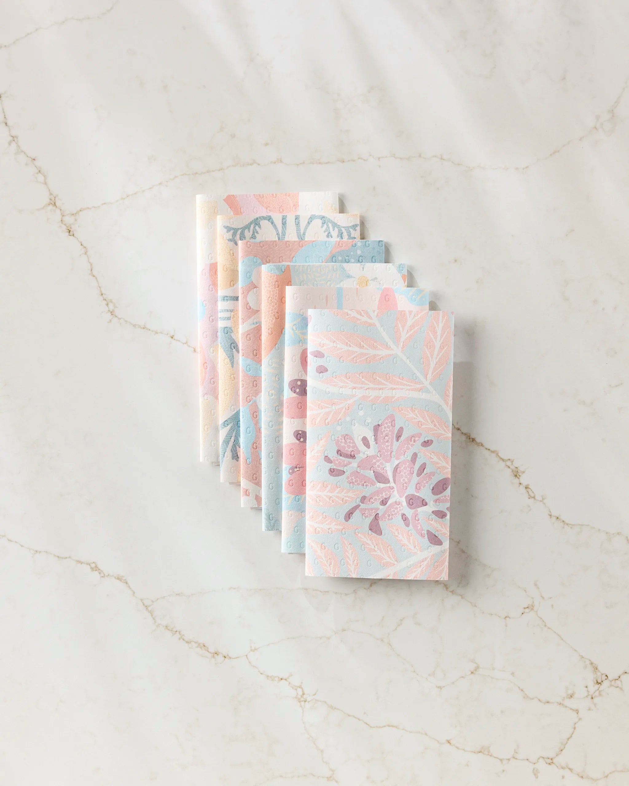 Seven State Flowers notebooks by Julianne Haness, featuring pastel floral designs, are arranged in a fan on a white marble surface next to reusable paper towels, creating an eco-friendly workspace.