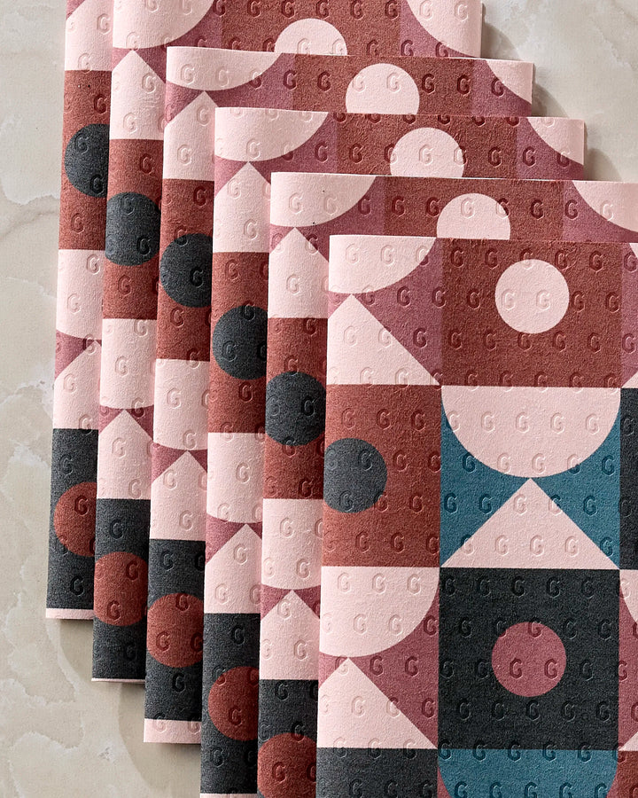 Five “Abstract Retro” notebooks by Jennifer Bouron, featuring geometric patterns in pink, red, blue, and black with subtle G designs, are stacked on marble—styled alongside reusable paper towels. Each cover showcases circles and triangles.