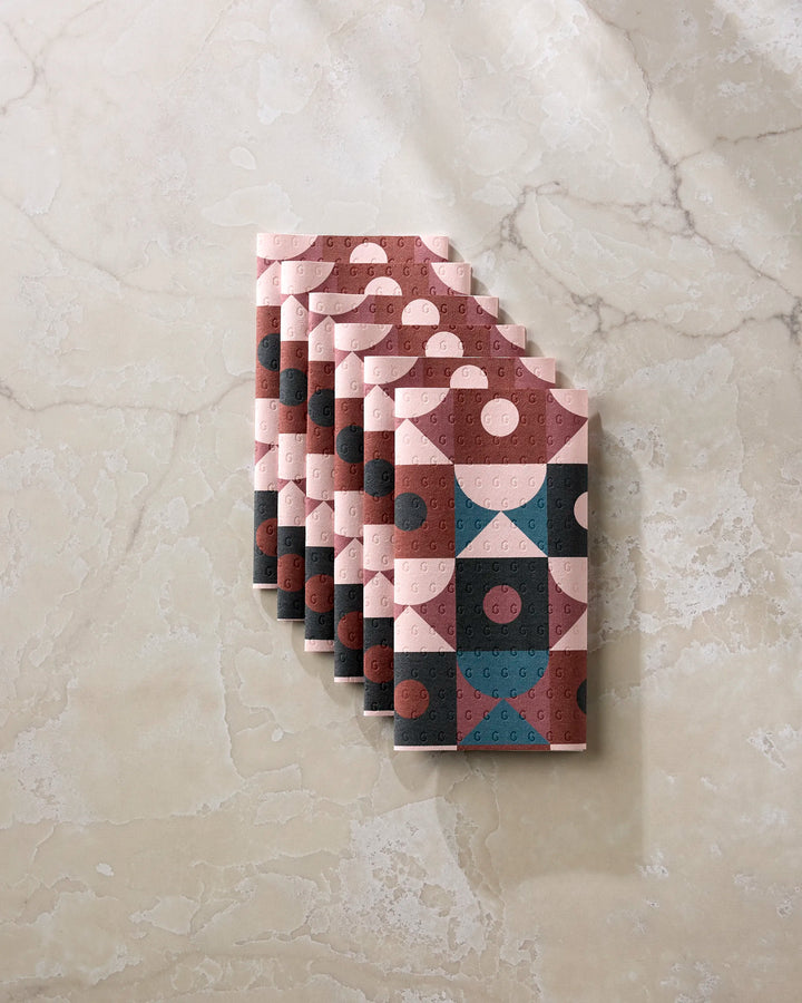 Six folded, reusable Abstract Retro paper towels by Jennifer Bouron, featuring a pink, teal, and brown geometric pattern, are stacked on a beige marble surface.