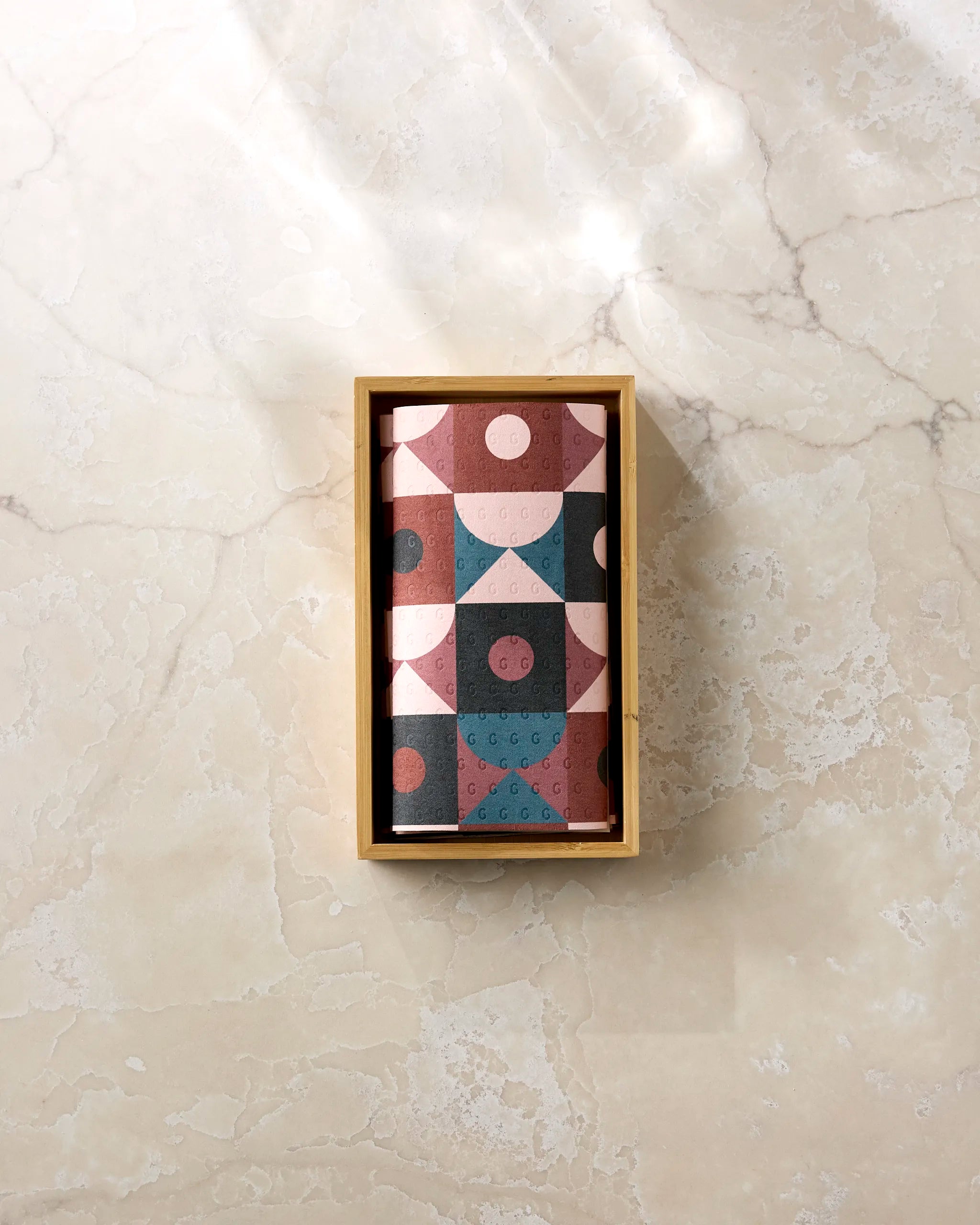 The Abstract Retro reusable paper towels by Jennifer Bouron, featuring pink, maroon, and teal geometric patterns, are displayed in a rectangular box on a light marble surface.