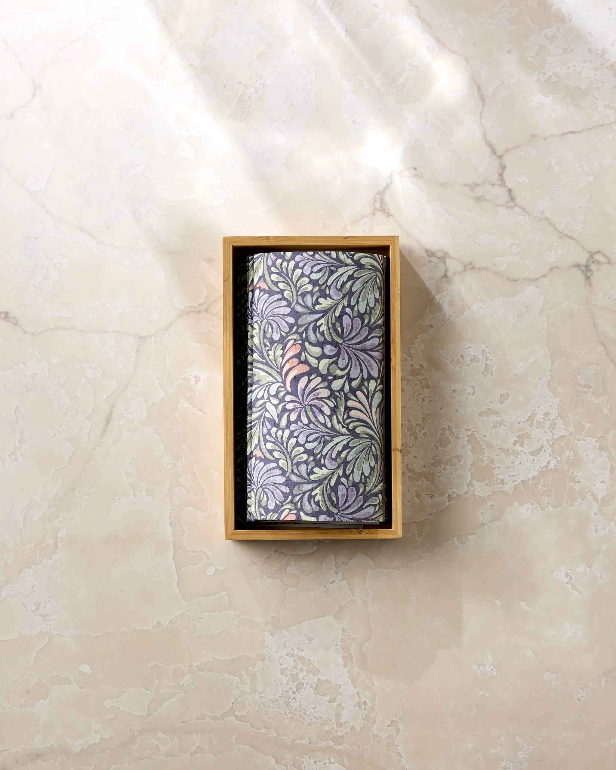 A rectangular box of Kris Ruff’s Acanthus eco-friendly kitchen towel, adorned with a decorative leafy pattern, rests on a light marble surface under gentle sunlight.