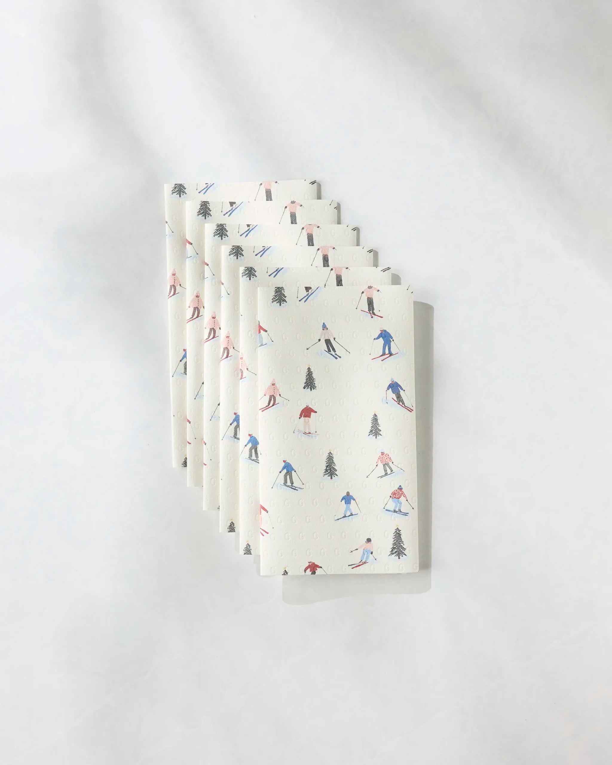 Six "Alpine Run" napkins by Geometry, featuring whimsical skiers and pine trees in red, blue, and black. This set of six eco-friendly napkins is a stylish alternative to paper towels for your table.