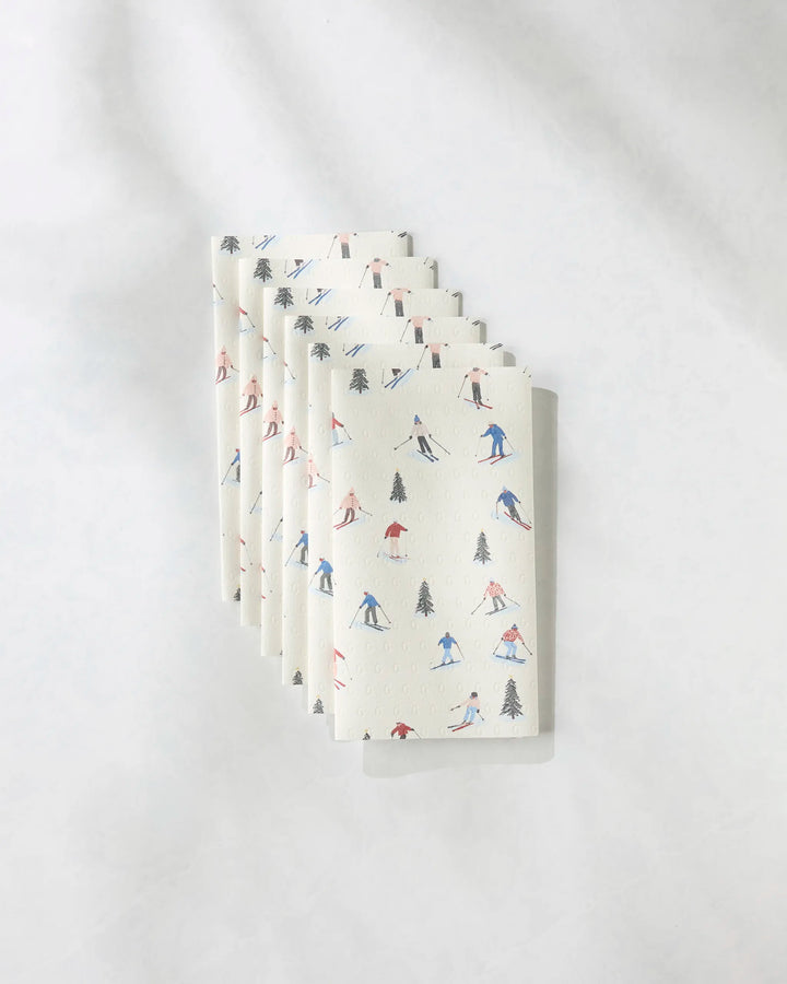 Six "Alpine Run" napkins by Geometry, featuring whimsical skiers and pine trees in red, blue, and black. This set of six eco-friendly napkins is a stylish alternative to paper towels for your table.