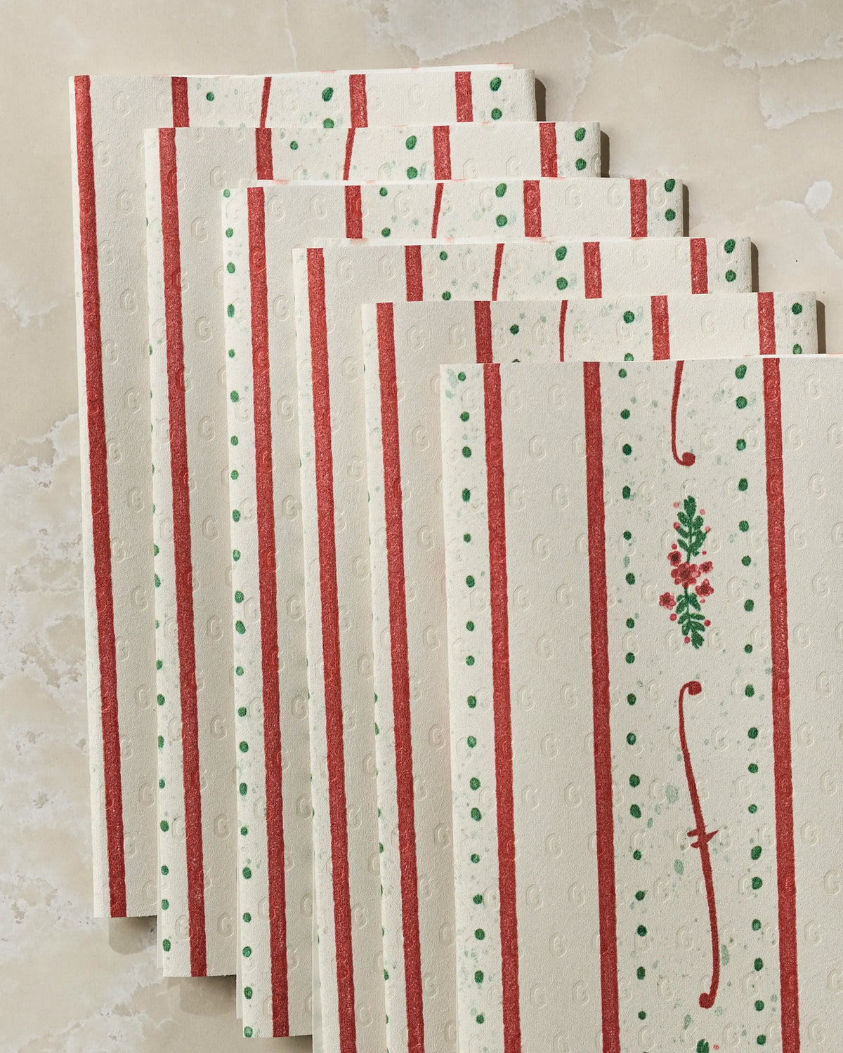 Six eco-friendly paper towels from Geometry’s Alpine Trim collection, featuring a cream-colored textured design with red stripes, green dots, and a small floral motif, are fanned out on a light marble surface.