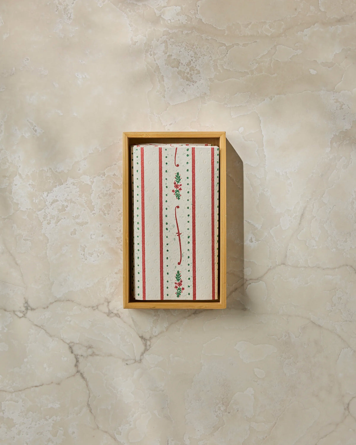A rectangular box from Geometry, called Alpine Trim, features a lid adorned with red, green, and white vertical stripes with small floral designs. Sitting on a beige marble surface, it’s perfect for storing eco-friendly paper towels or other recycled paper products.