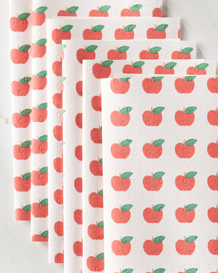 Brittany Jepsen’s Apple Grove features six white reusable paper towels stacked in a staggered pile, each adorned with a repeating red apple and green leaf pattern on a light, neutral surface.