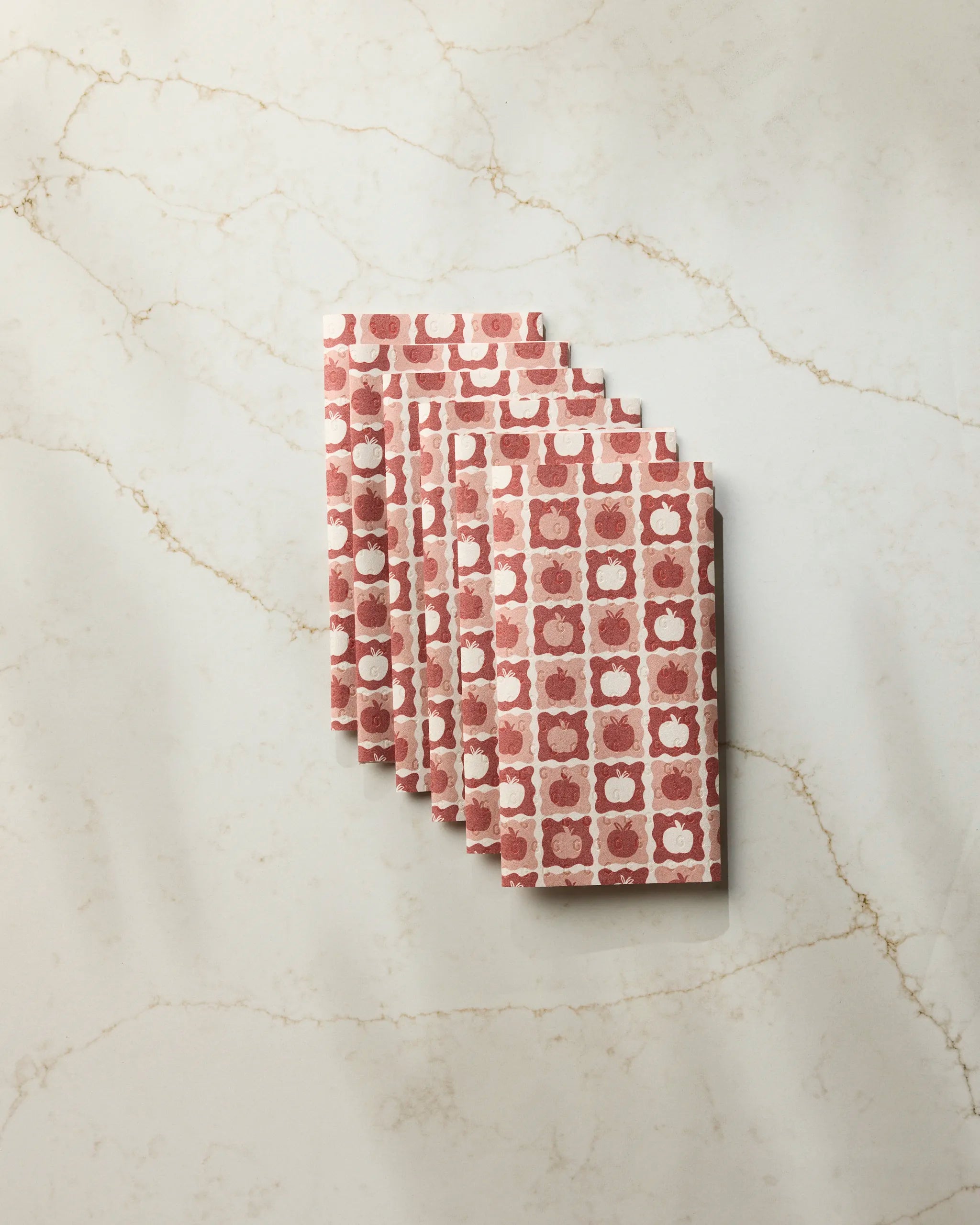 Five Apple Quilt patterned red and white eco-friendly kitchen towels by Geometry are fanned out on a white marble surface with beige veining.