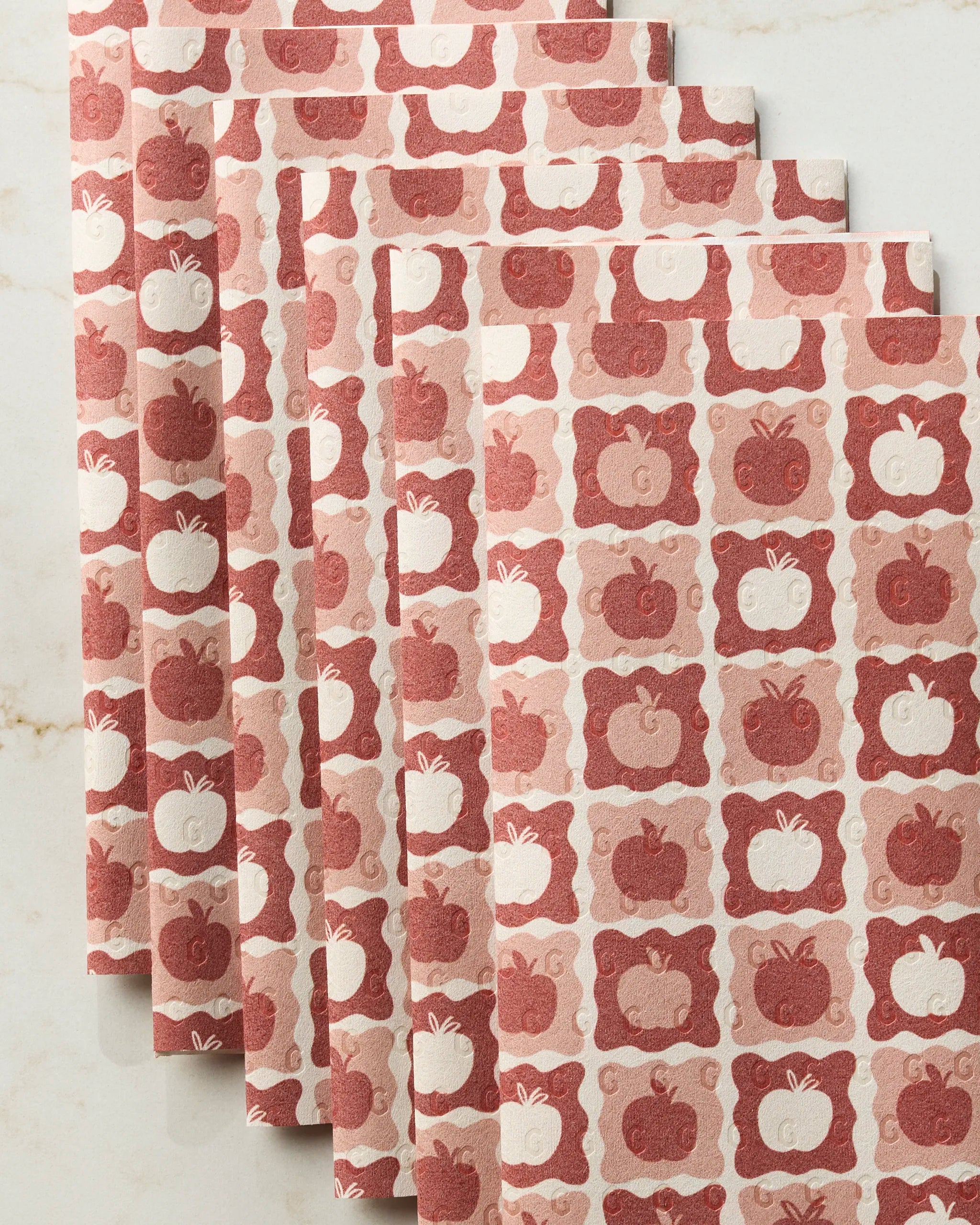 Five folded, reusable Apple Quilt paper towels from Geometry, featuring red and white apple and “G” patterns, are fanned out on light marble—an eco-friendly kitchen towel choice.