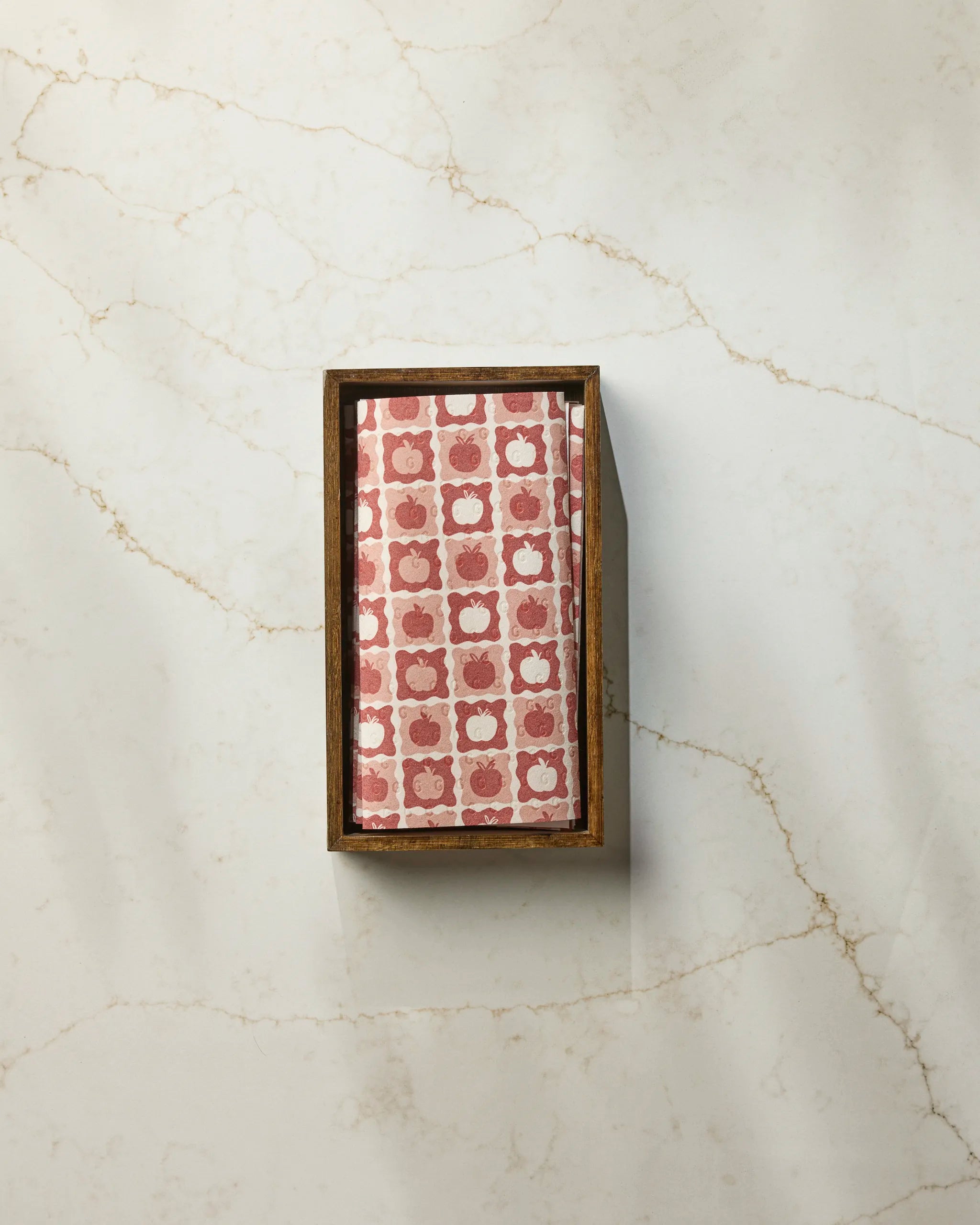 The Geometry Apple Quilt is a rectangular wooden tray featuring a neatly folded, pink and white patterned napkin, styled like eco-friendly kitchen towels, resting on a white marble surface with subtle brown veining.