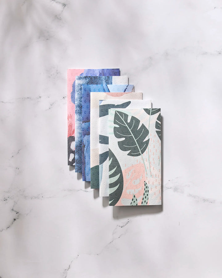 Five folded GEOMETRY Artistic dishcloths, featuring botanical and abstract designs in green, pink, blue, and white, are arranged on white marble—a sustainable alternative to disposable options.