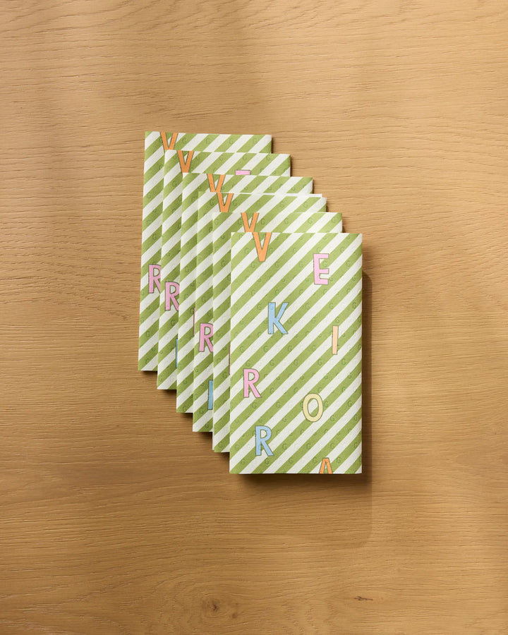 Six eco-friendly paper towels from Geometry, featuring green and white diagonal stripes and colorful scattered letters, are fanned out on a wooden surface.