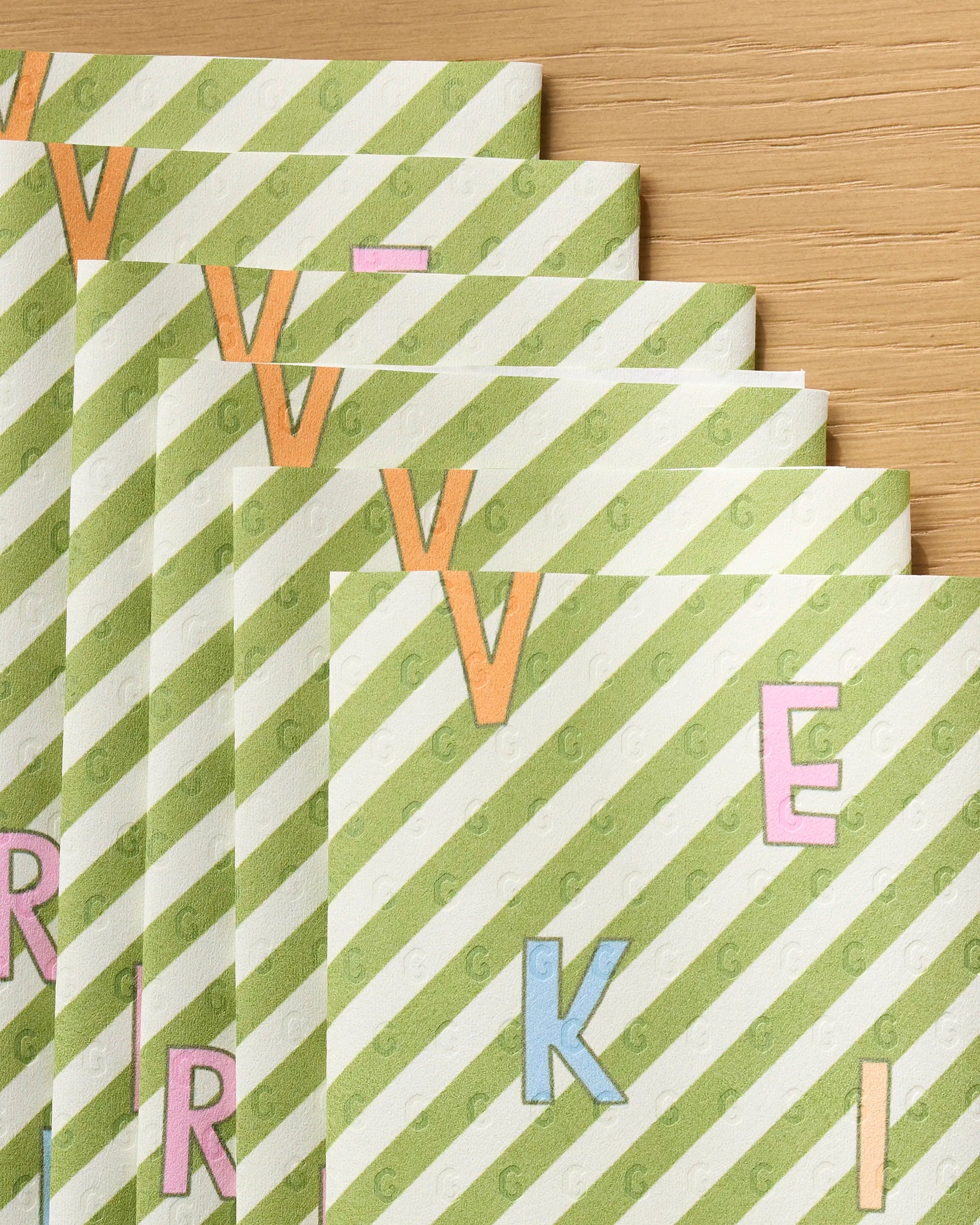 Several recycled paper towel sheets from Geometry’s Attributes collection, featuring diagonal green and white stripes and colorful letters like V, E, R, and K, are fanned out on a wooden surface.